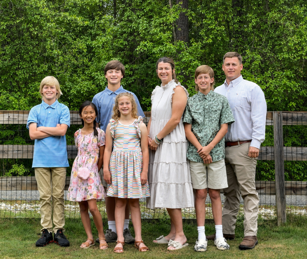 A family of seven standing outdoors in front of a wooden fence and green trees, smiling for a photo. The family includes two adults and five children, dressed casually.