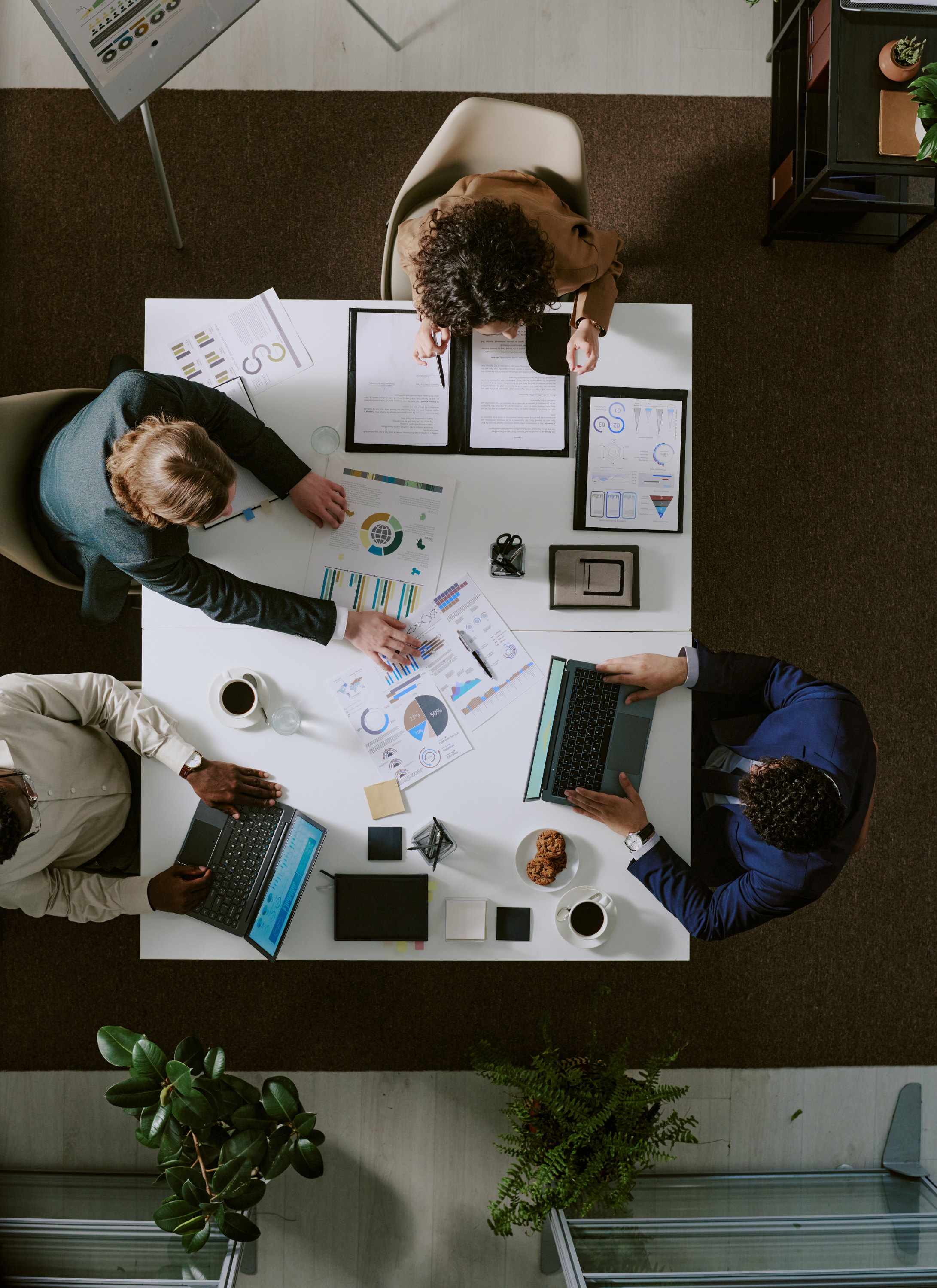A top-down view of a diverse group of five people working together in a meeting room. They are seated around a white table with laptops, printed charts, documents, coffee cups, and snacks.
