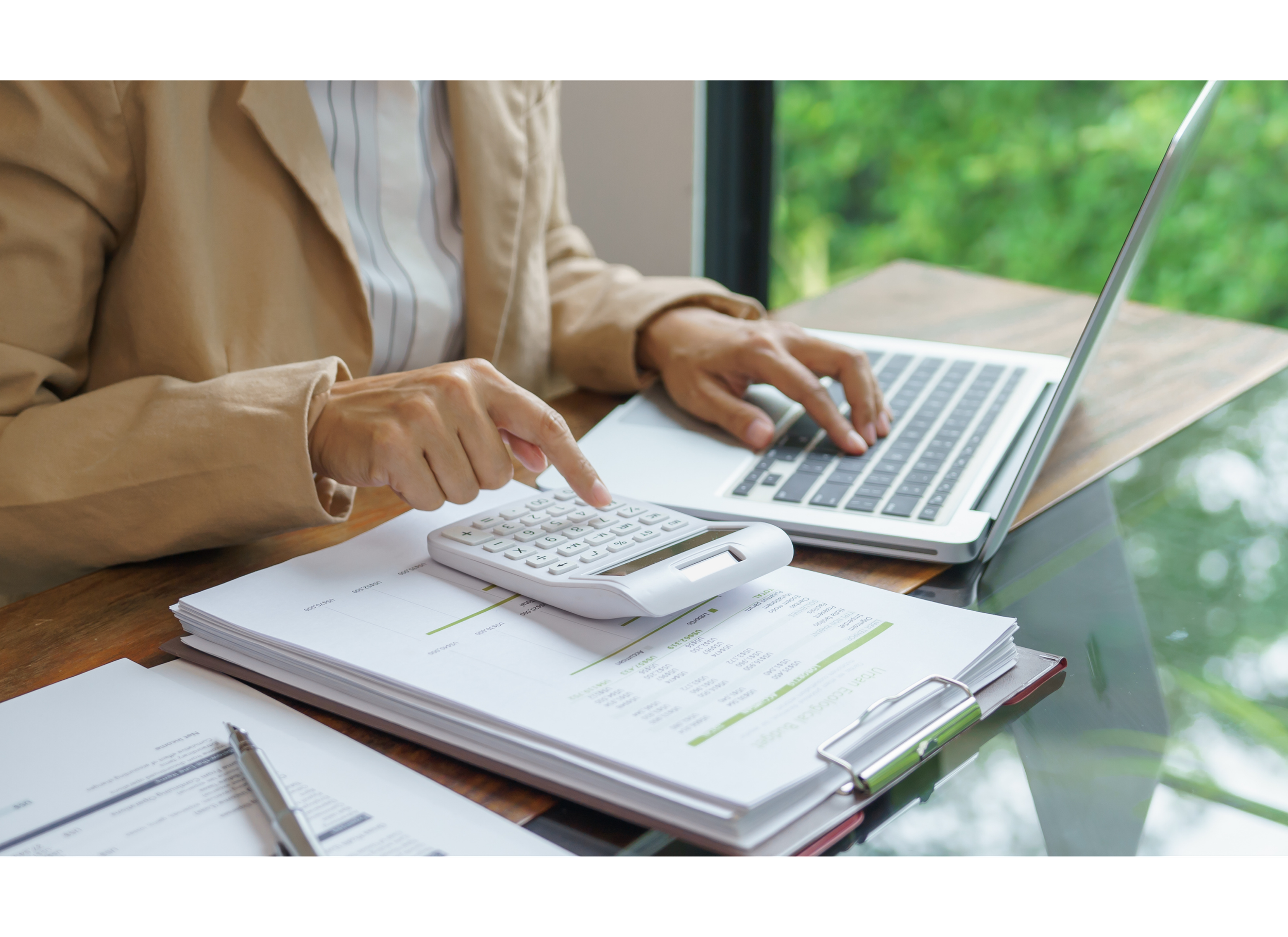 Person working with a calculator and laptop on a wooden desk, surrounded by documents, with a view of greenery outside a window.