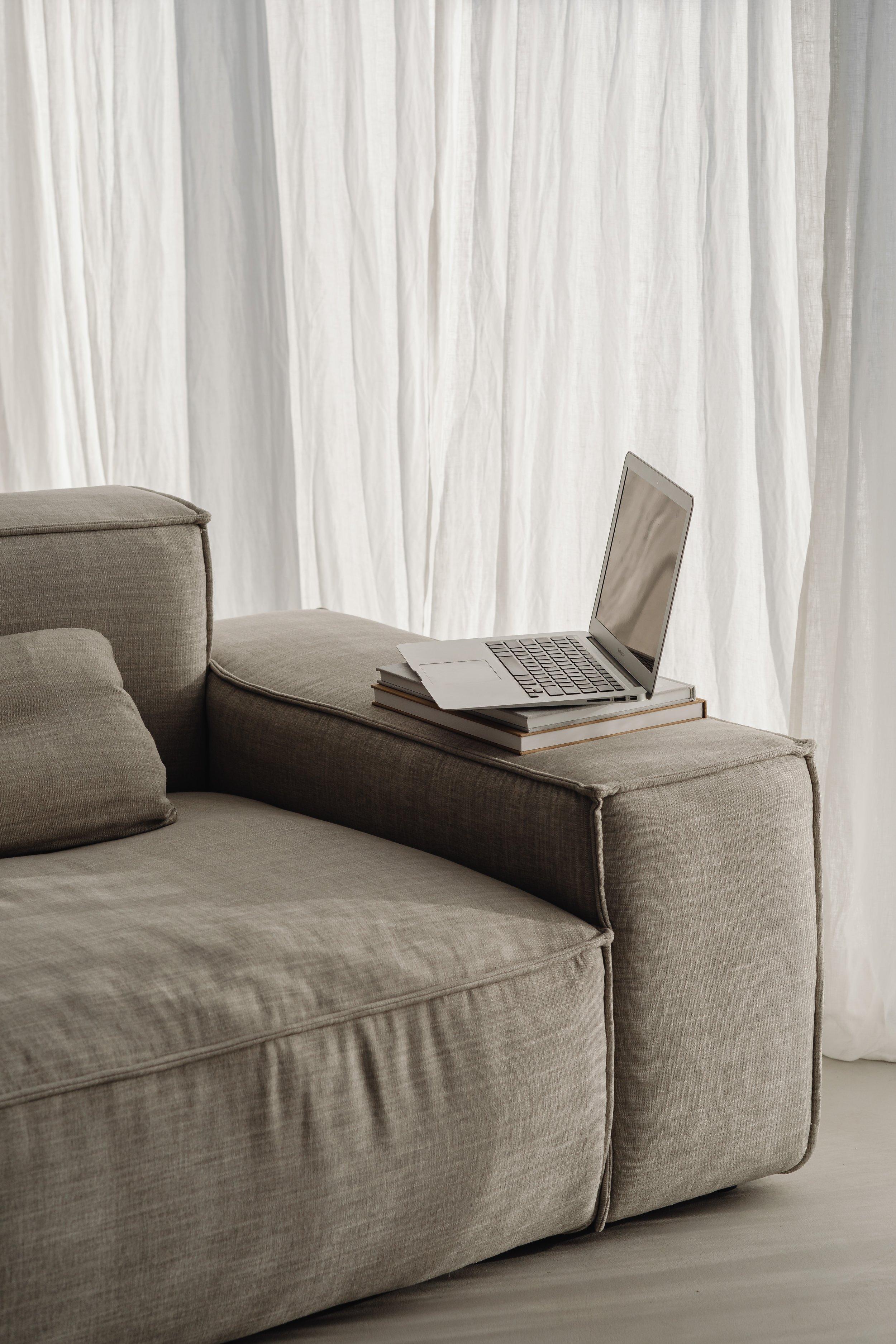 A beige sectional sofa with cushions in front of a white curtain, with an open laptop and a stack of books on the armrest.