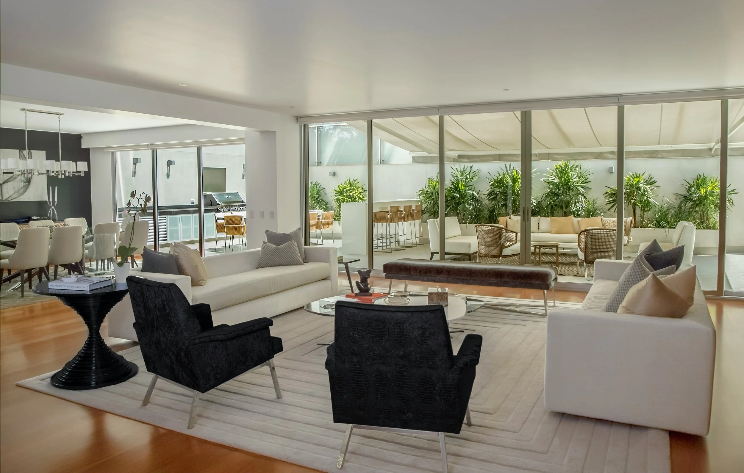 A modern living room with white sofas, black and dark gray chairs, a glass top coffee table, and an area rug. There are large floor-to-ceiling windows with plants outside and a dining table with white chairs visible in the background.