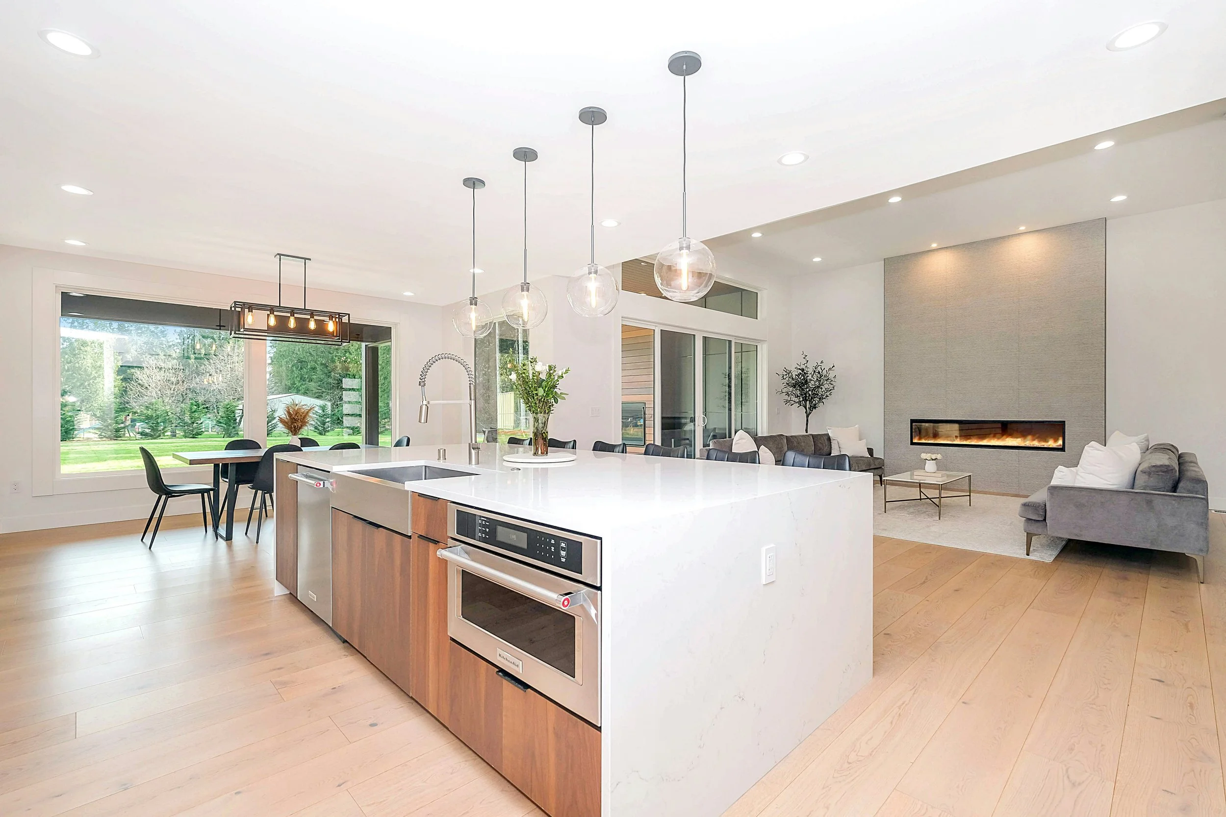 Open-concept kitchen and living room with a large white island, modern hanging light fixtures, a dining table, large windows, and a fireplace in the background.