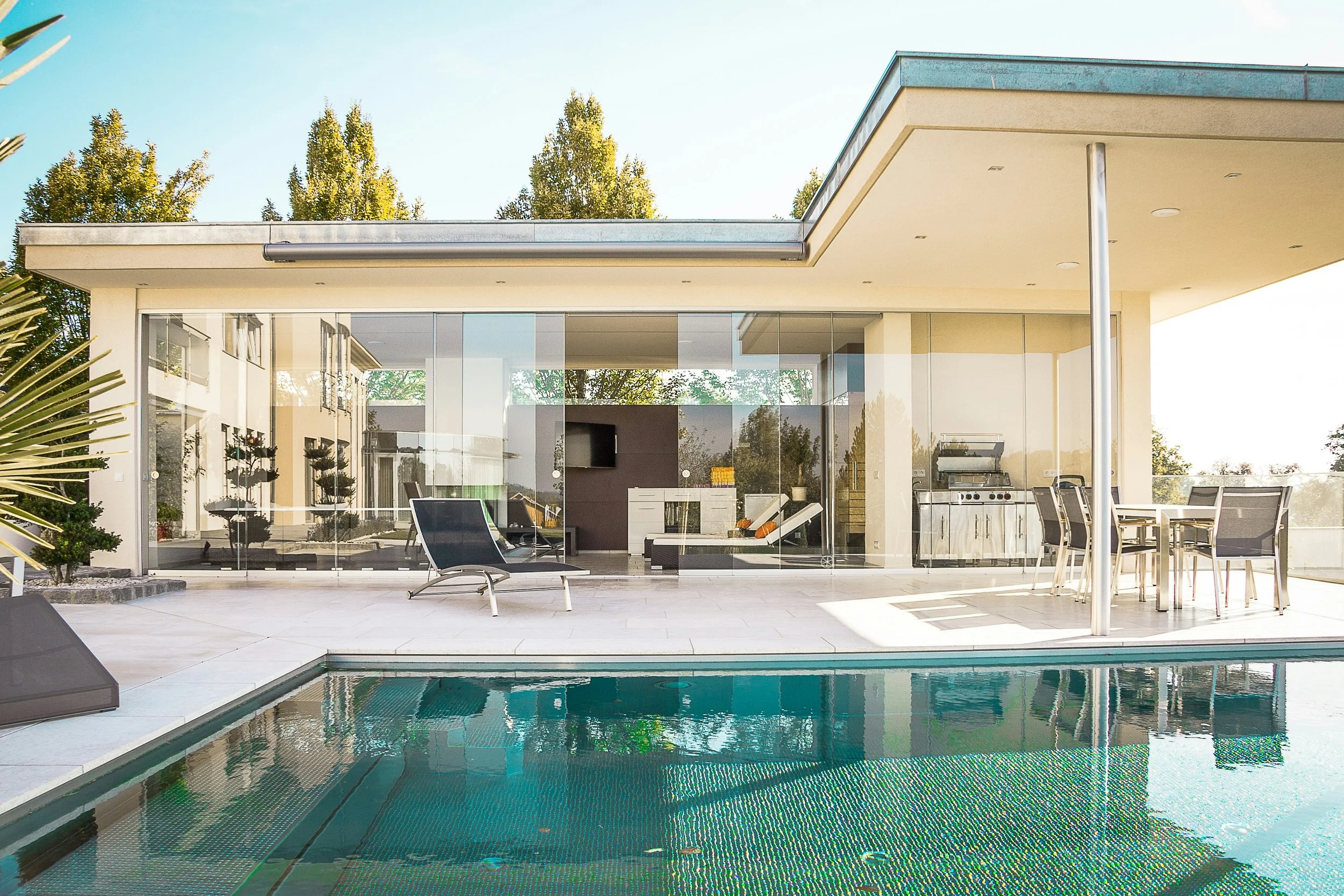 Modern house with a glass wall, outdoor seating and grill area, and a swimming pool in front, surrounded by trees on a sunny day.