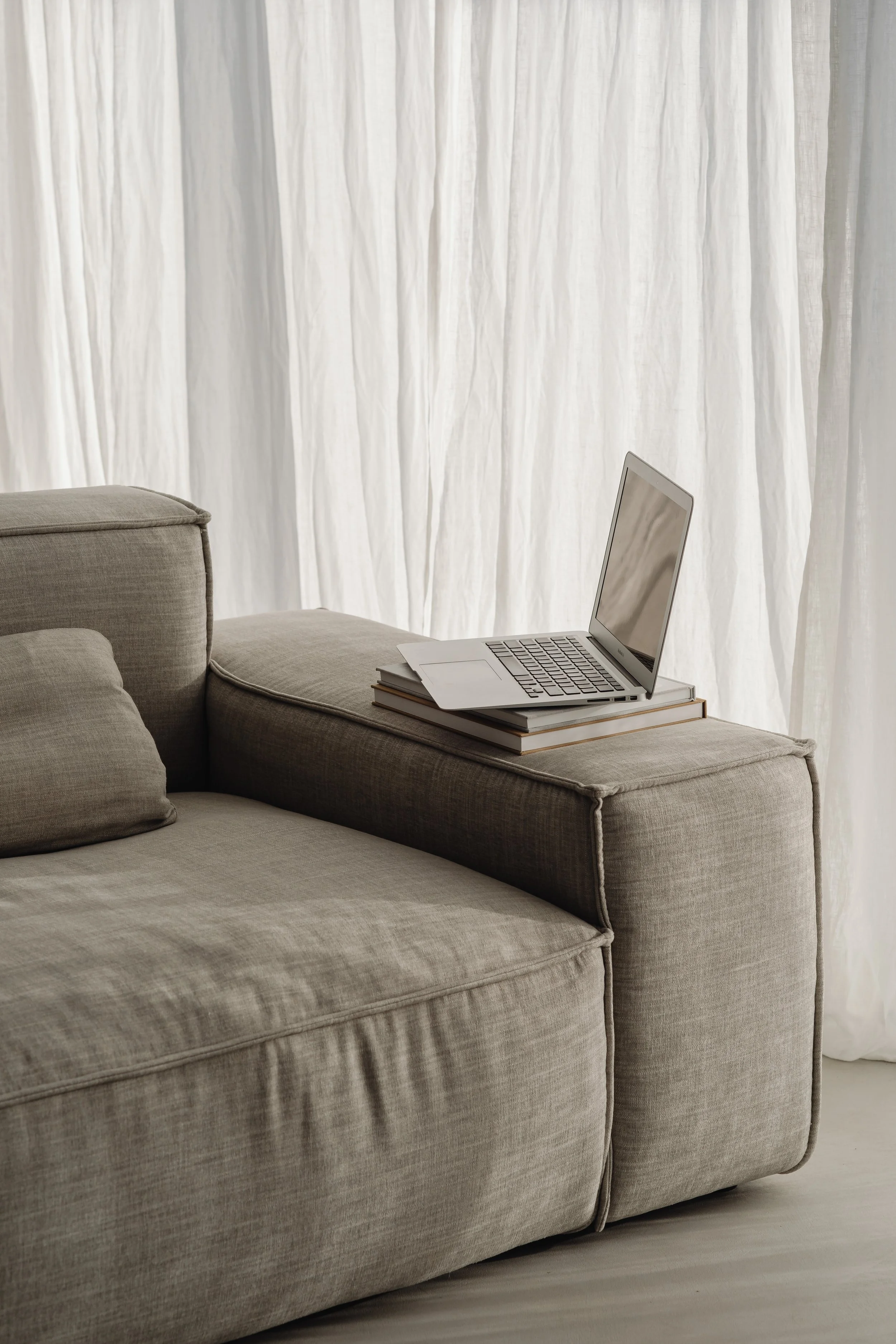 A beige upholstered sofa with a laptop and a closed book on the armrest, in front of white curtains.