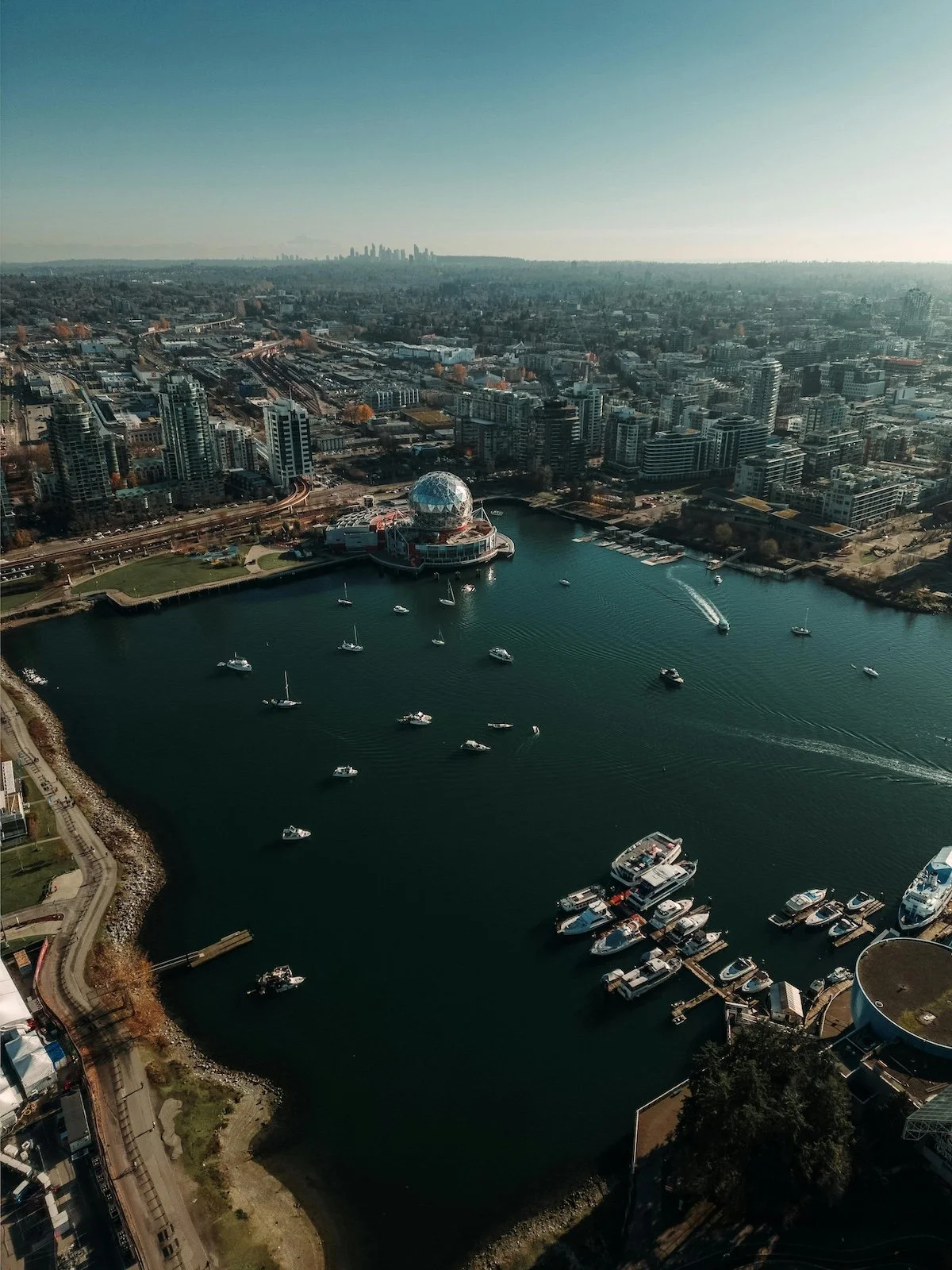 Aerial view of a city with a water body in the foreground, featuring boats and yachts, with a cityscape and skyscrapers in the background.
