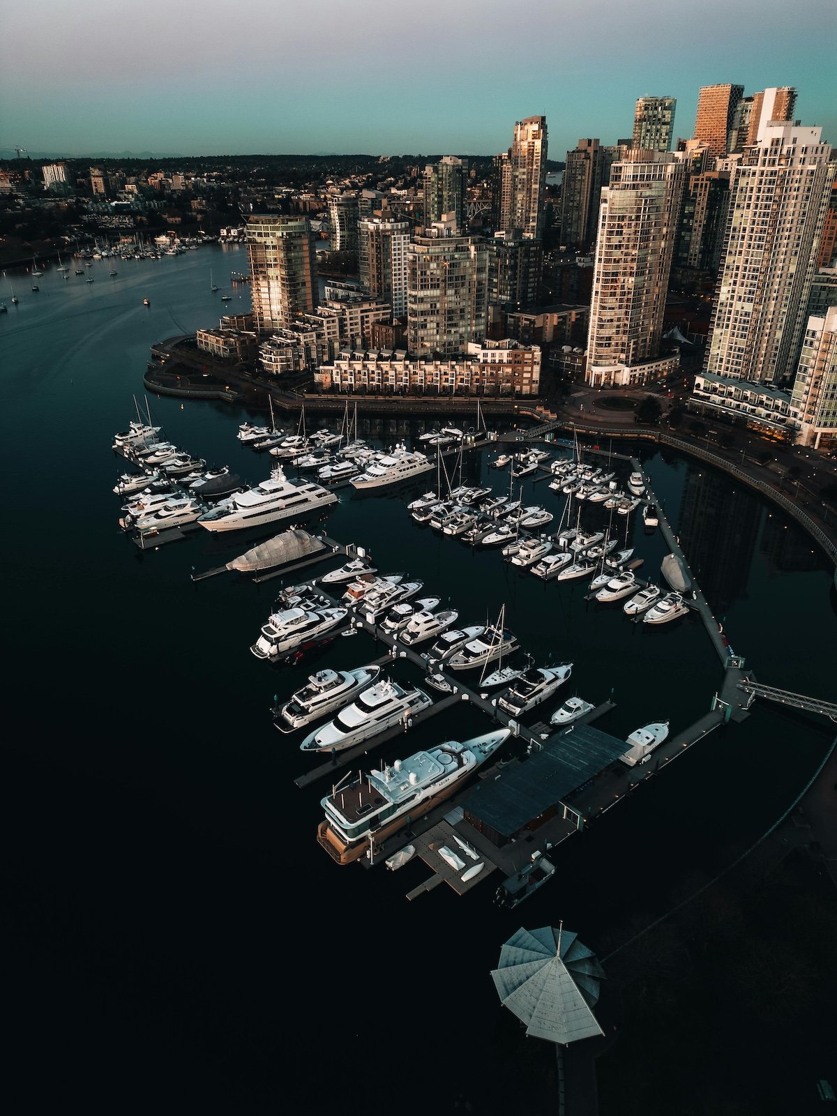 Aerial view of a city marina with numerous luxury yachts docked, high-rise buildings in the background, and an umbrella over a small structure on the dock.