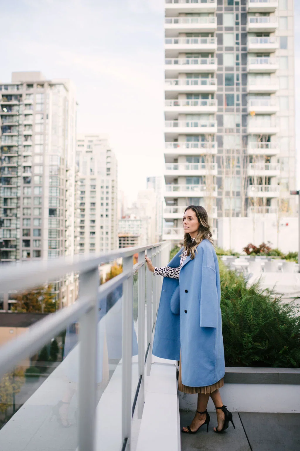 A woman in a blue coat and high heels standing on a balcony with city buildings in the background.