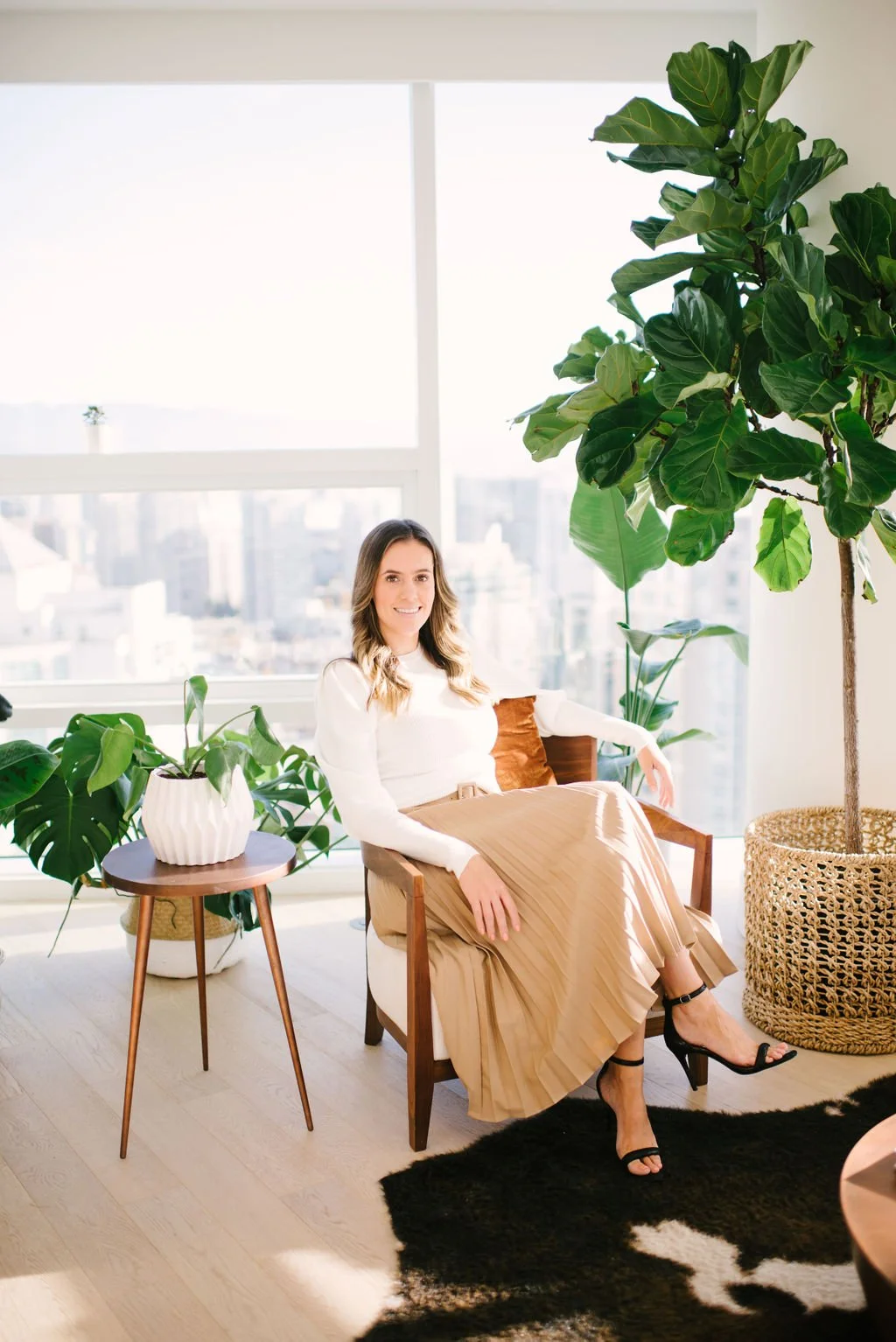 A woman sitting in a modern, sunlit apartment with large windows, surrounded by green houseplants, including a tall fiddle leaf fig and a monstera, with a city skyline visible outside.