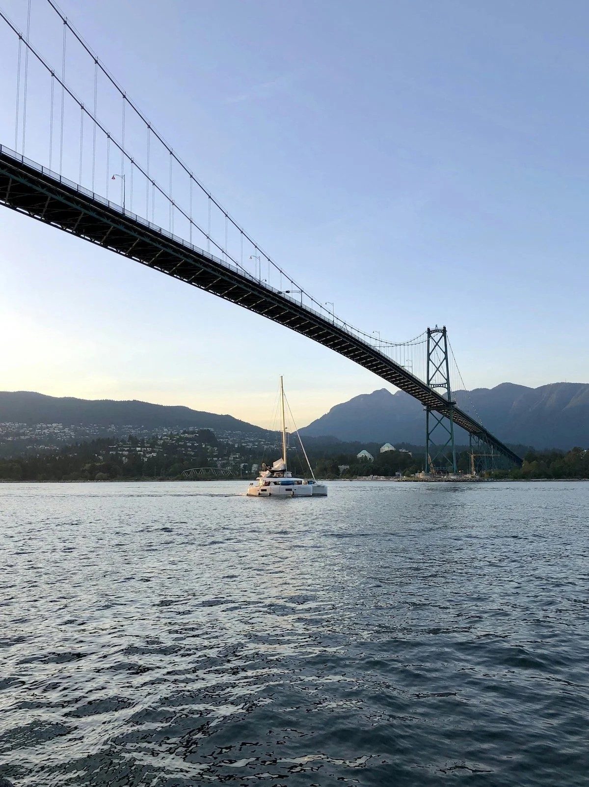 A sailboat on a calm body of water beneath a large suspension bridge, with mountains in the background, during early evening or sunset.