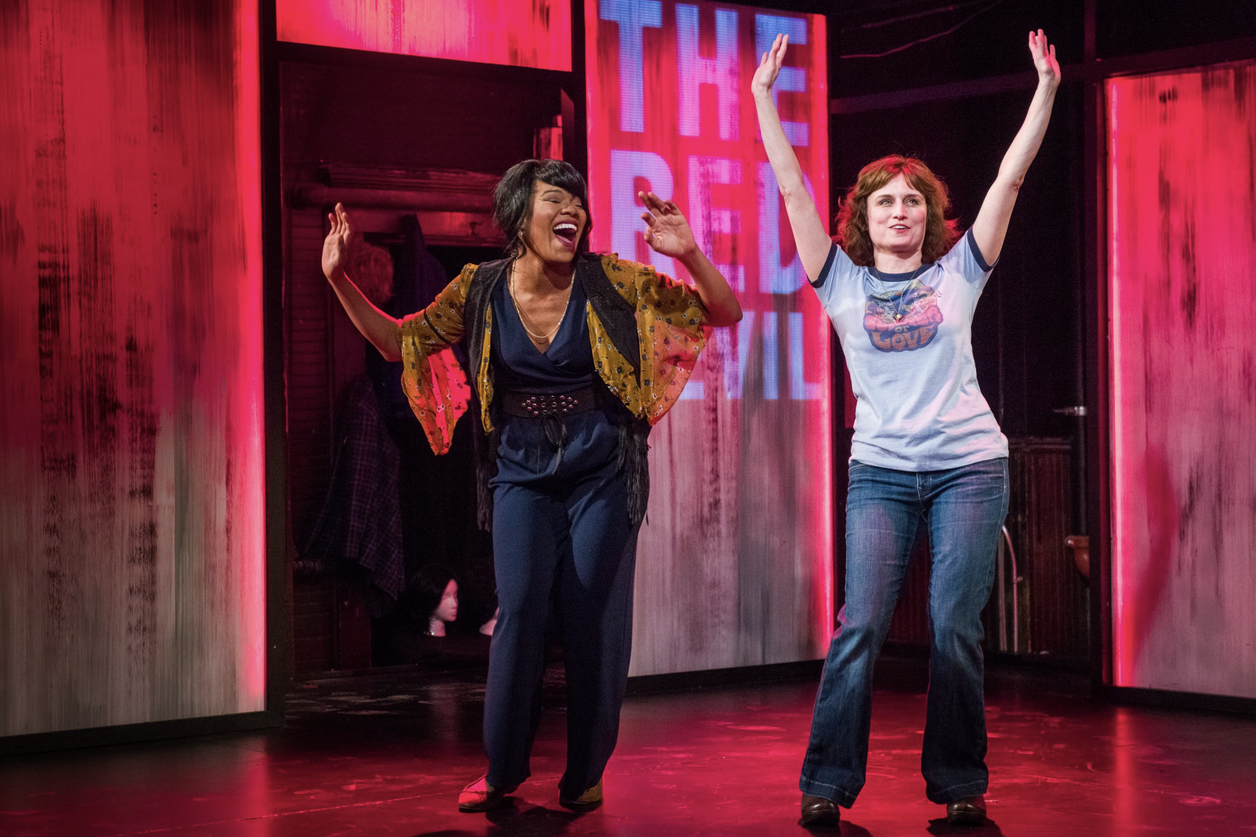 Two women dancing and smiling on stage with pink lighting and a neon sign in the background.