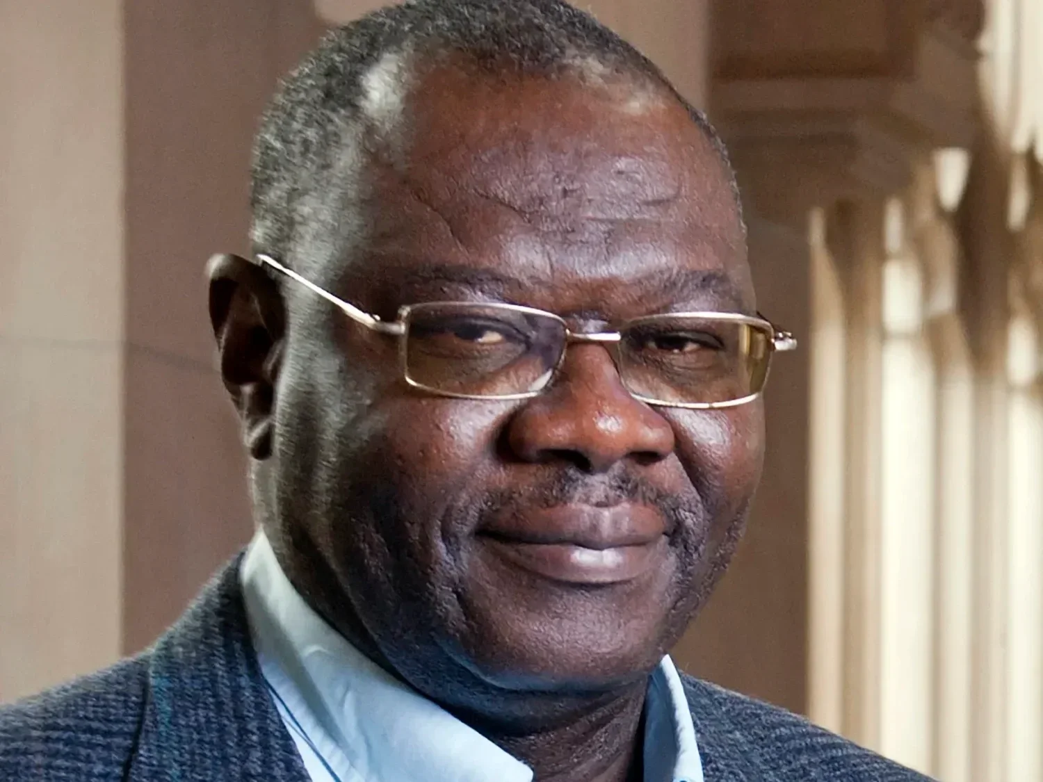 Close-up portrait of an older African American man with glasses, wearing a suit jacket and in front of a window with curtains.