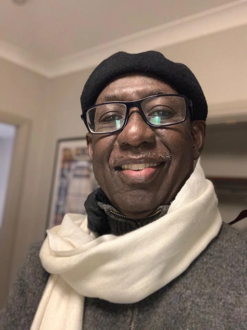 A smiling man wearing a black beret, glasses, a dark sweater, and a cream-colored scarf in a cozy indoor setting.
