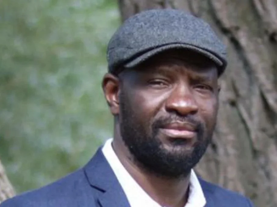 Close-up of a man with a beard wearing a gray flat cap and a navy blazer outdoors near a tree.