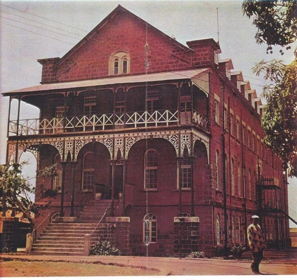 A large, historic, red brick mansion with a wraparound porch, ornate woodwork, and multiple stories, situated outdoors with a person walking nearby.