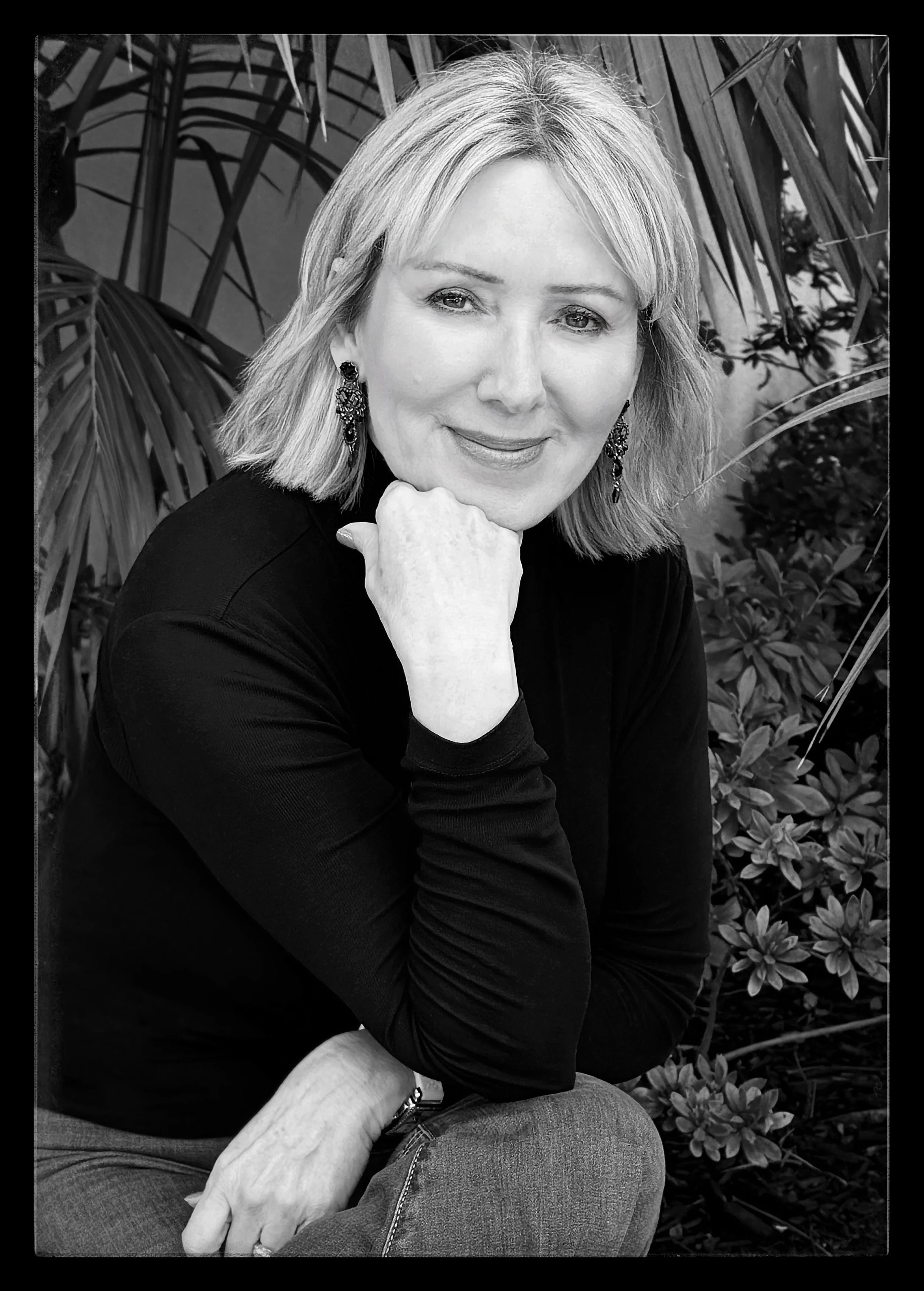 A black and white photograph of a woman with shoulder-length hair, wearing earrings and a long-sleeved top, sitting outdoors amidst foliage, with her chin resting on her hand and smiling gently.