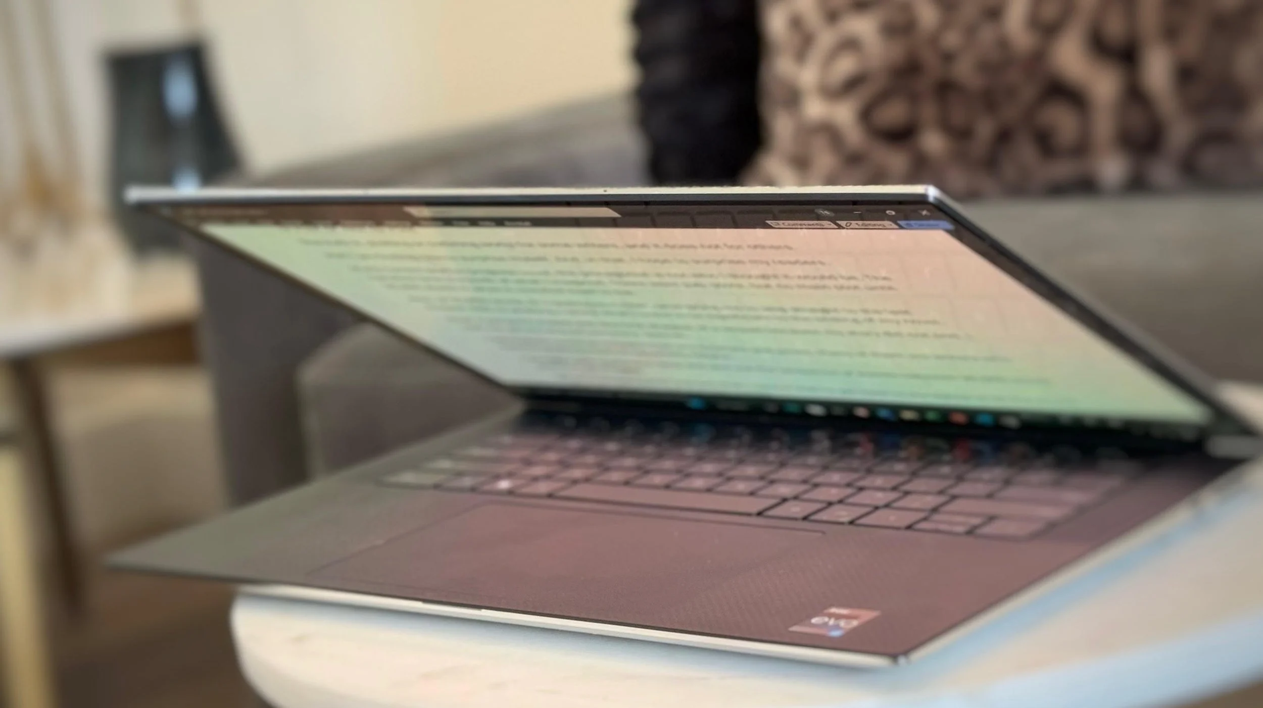 A close-up of a laptop with the screen tilted back, showing a blurred document or webpage, placed on a gray surface with a plant or decorative object in the background.