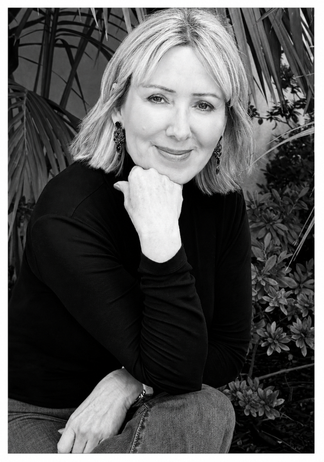 A black and white portrait of a woman with blonde hair, wearing earrings, sitting outdoors among plants, smiling and resting her chin on her hand.
