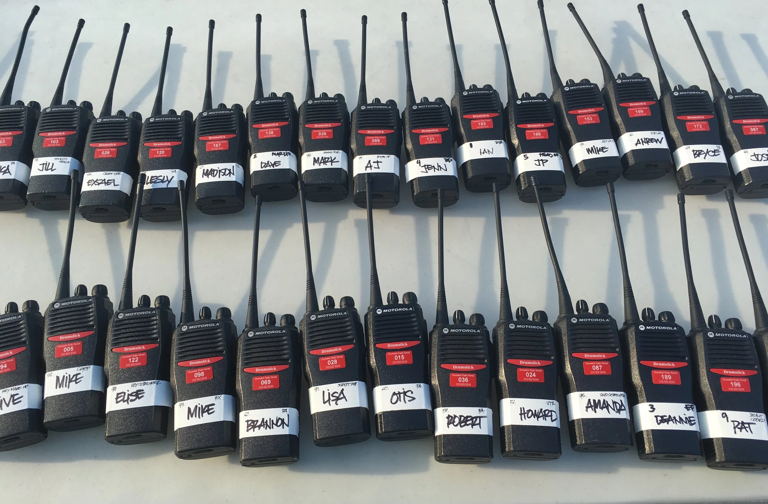 Multiple Motorola two-way radios lined up on a table, each with a white label bearing a handwritten name or initials.