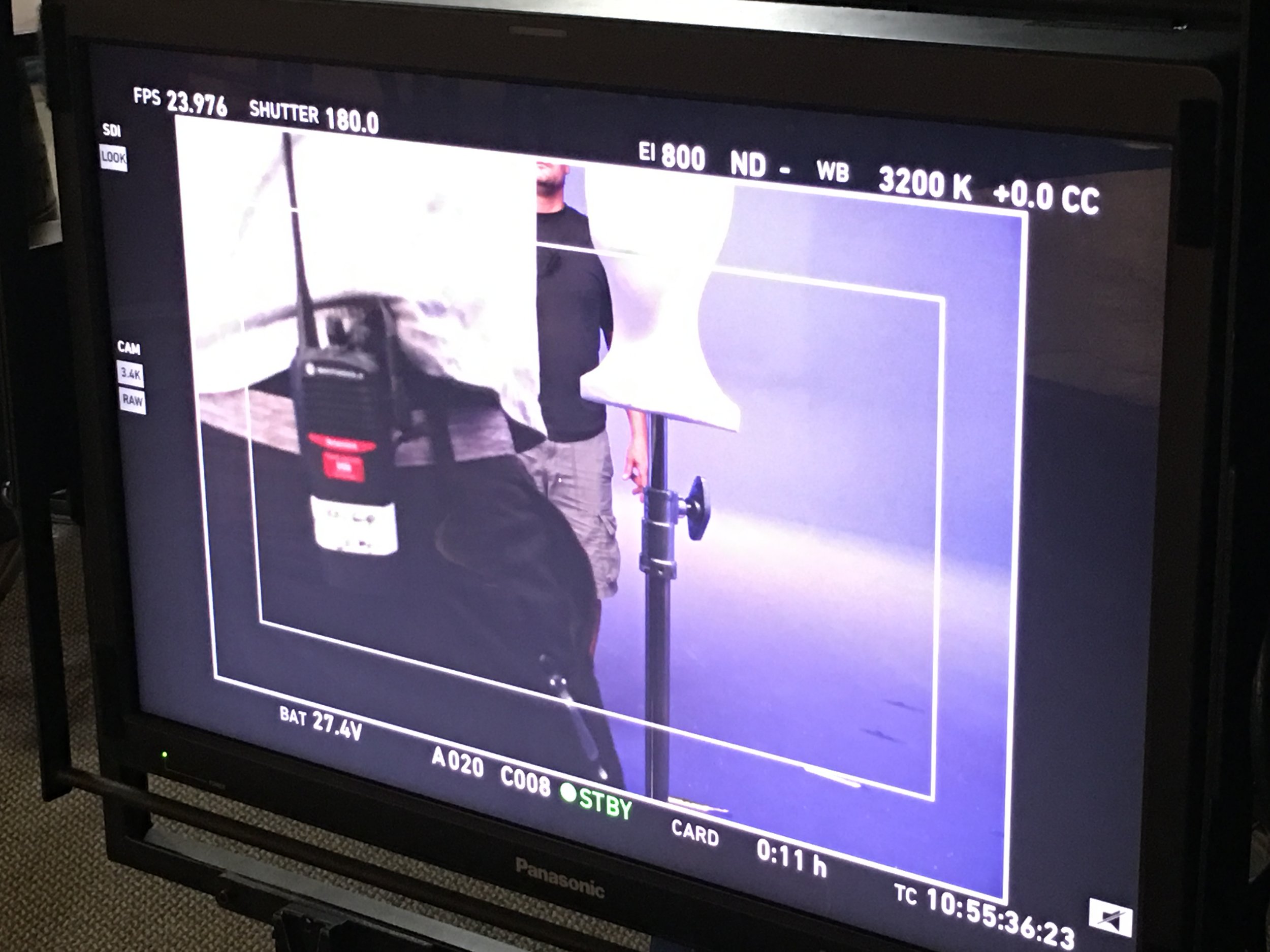 on a film or television set, a person's silhouette is visible in the background, with various equipment and a white reflector or backdrop in the foreground.