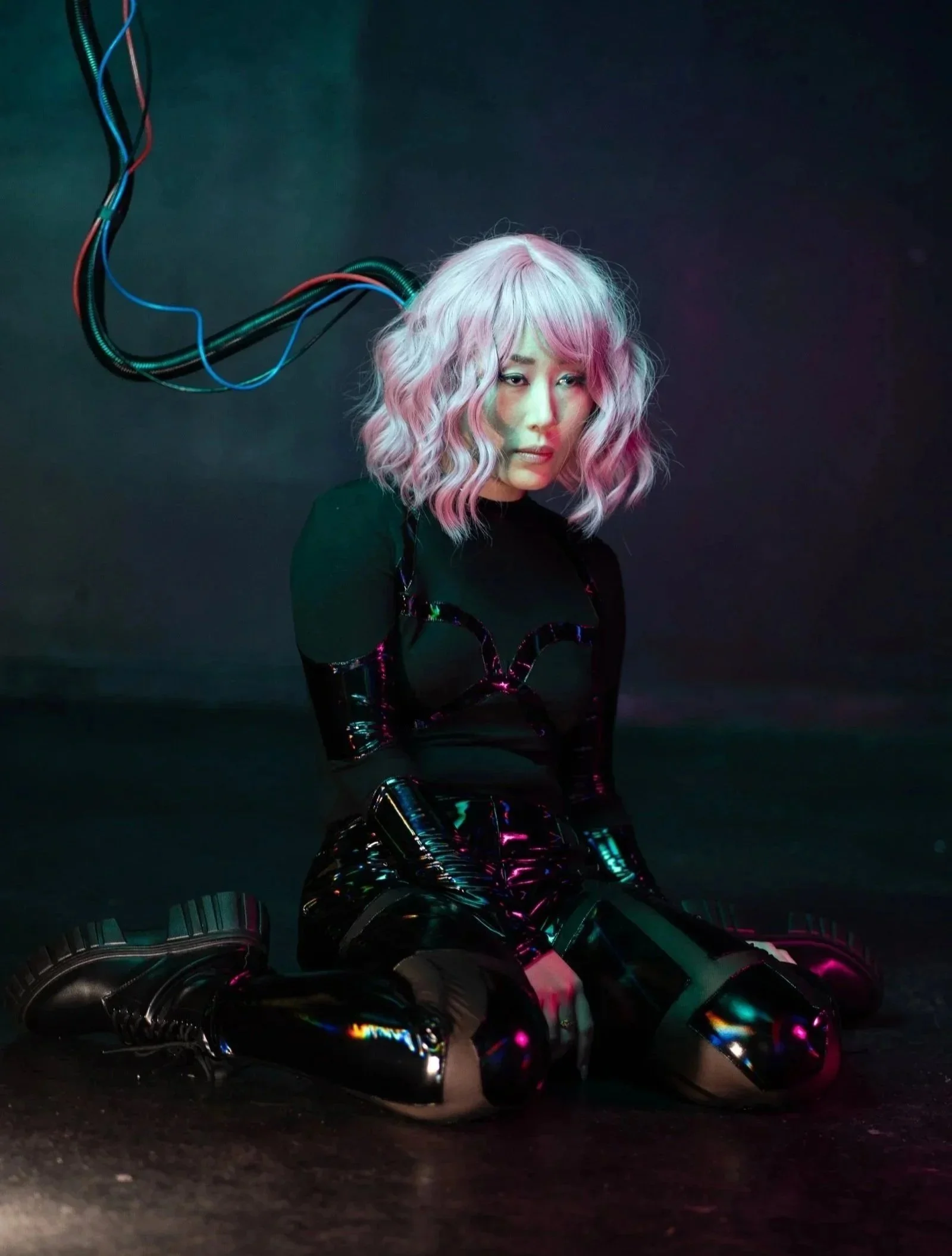 A woman with pastel pink and silver wavy hair in a dark setting with colorful lighting. She is wearing black shiny boots and a black shiny outfit, sitting on the ground with her legs extended.