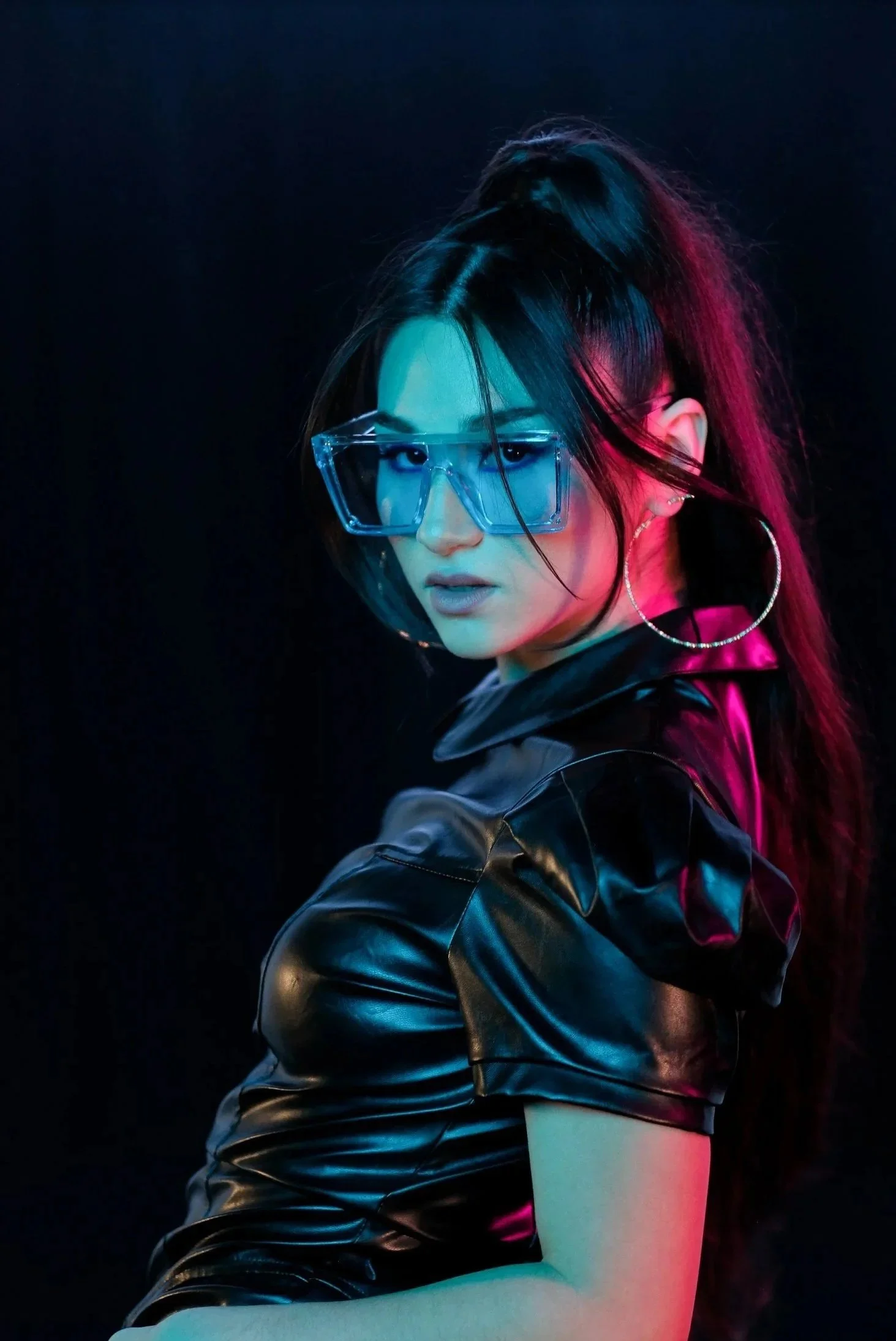Woman with long dark hair tied in a high ponytail wearing oversized clear glasses, large hoop earrings, and a shiny black leather jacket posed against a dark background with blue and pink lighting.