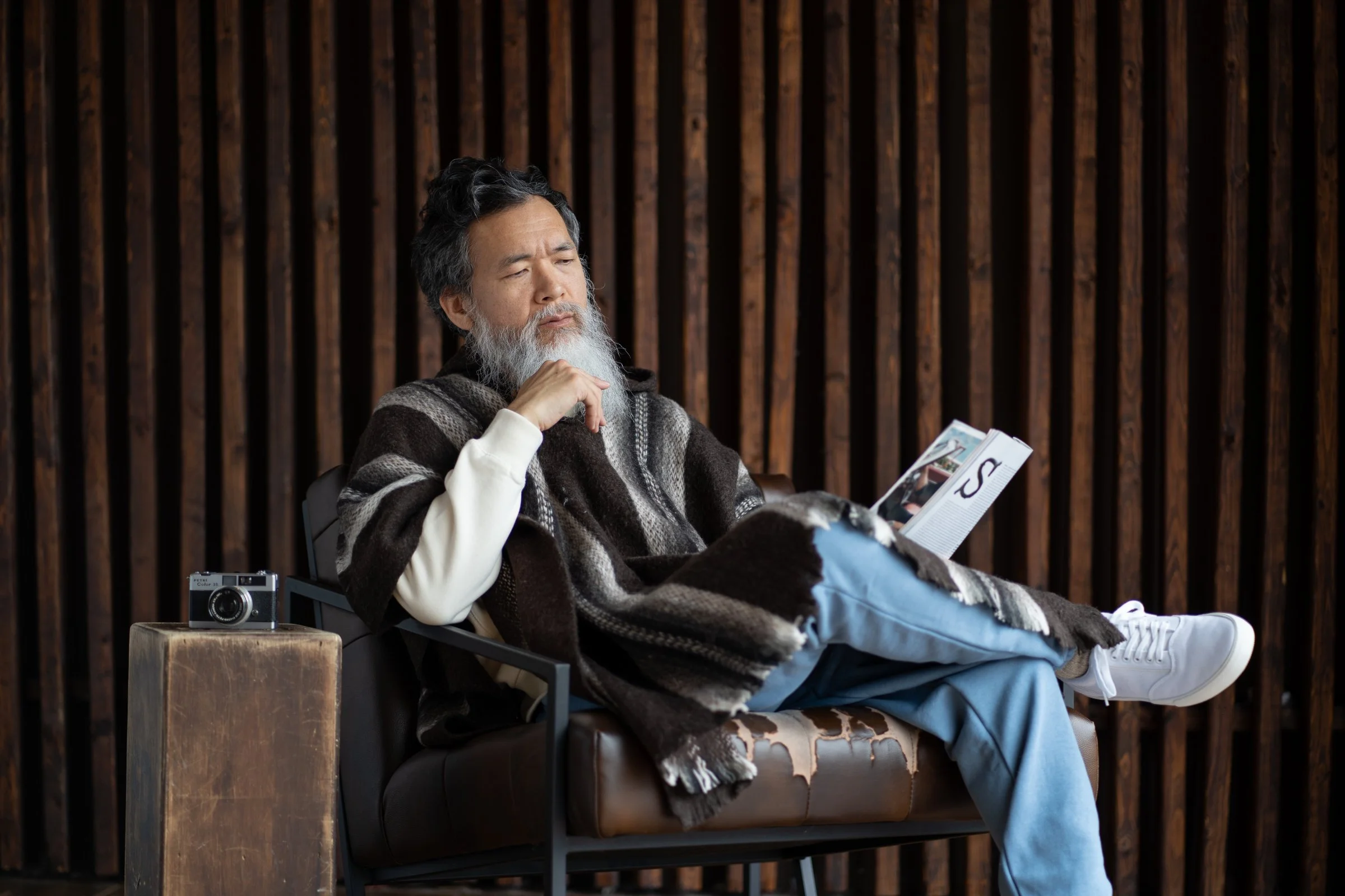 Bearded man sitting on a leather chair, reading a magazine, with a camera placed on a wooden side table next to him, against a dark wooden plank wall background.