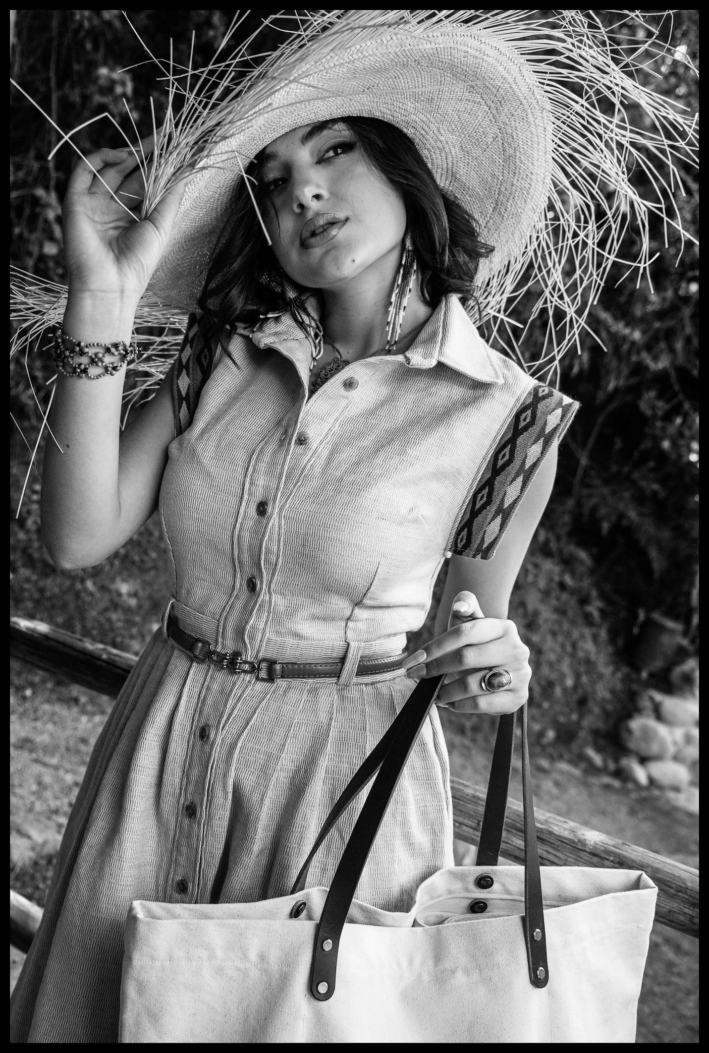 A woman with dark hair wearing a large sun hat, a sleeveless button-up dress with a patterned sleeve detail, and a bracelet on her right wrist, standing outdoors and holding a tote bag with leather straps.