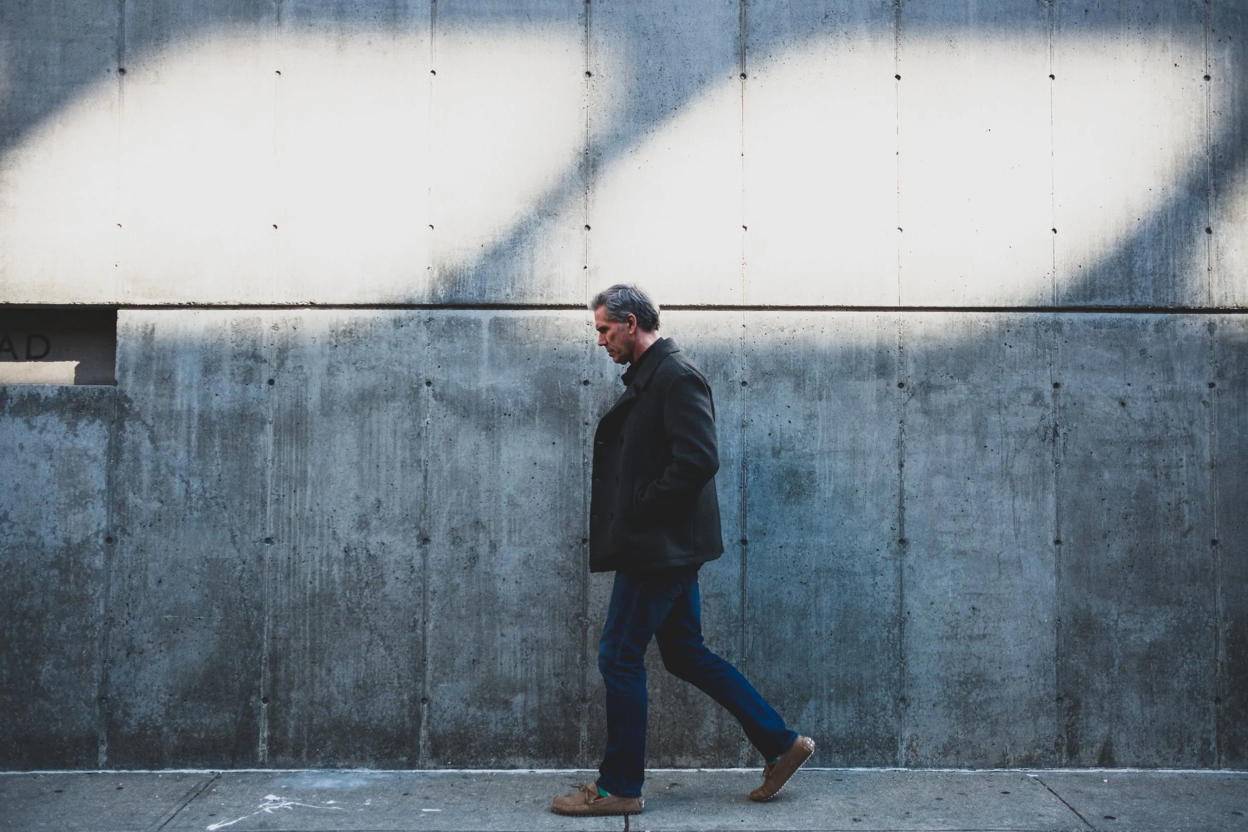 A man walking past a concrete wall with shadow and texture, wearing a black coat, blue jeans, and tan shoes.