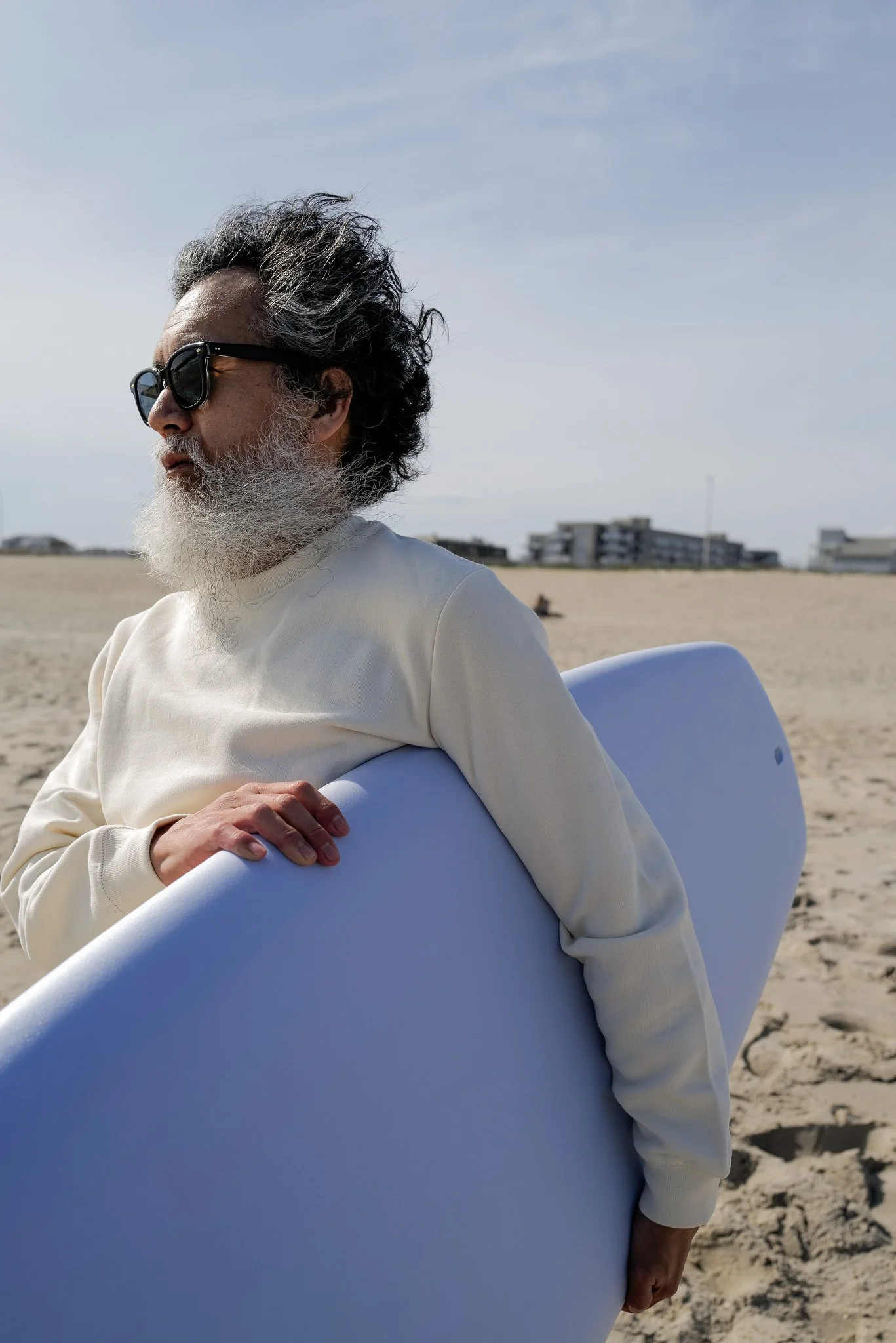 An older man with gray hair and a beard, wearing black sunglasses and a white long-sleeve shirt, holding a surfboard on a sandy beach with buildings in the background.