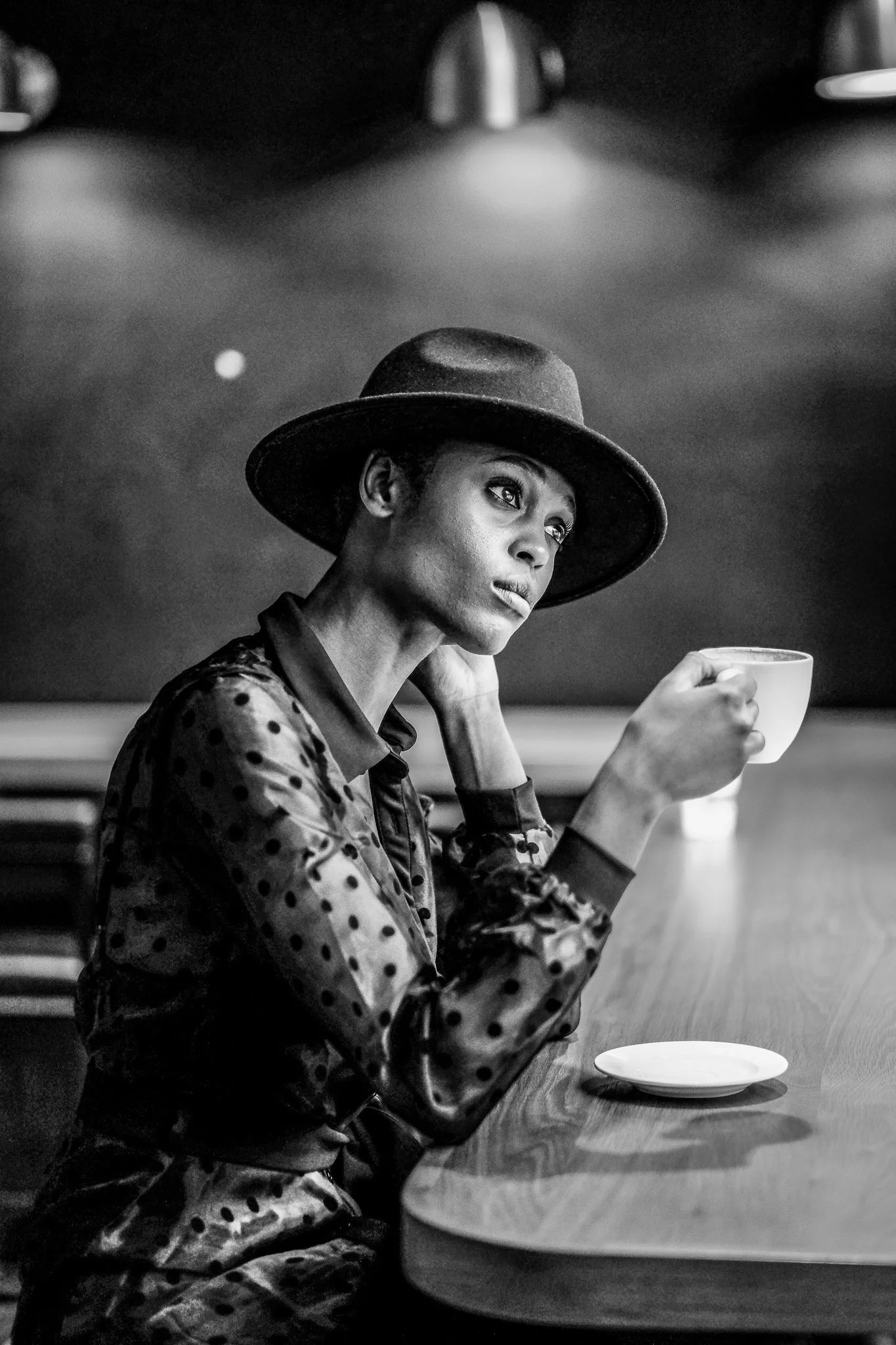 A woman with short hair, wearing a wide-brimmed hat and a polka-dot blouse, sitting at a table with a coffee cup in her hand in a dimly lit setting.