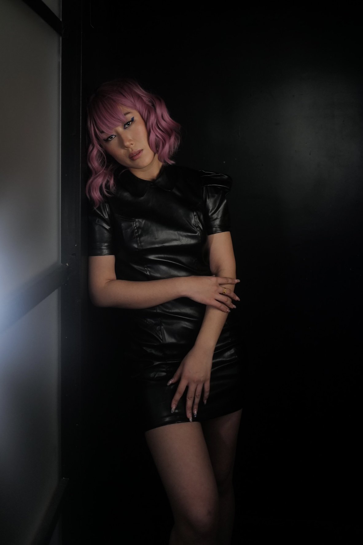 A woman with wavy pink hair and makeup, wearing a black leather dress, standing in a dark corner with black walls, posing with her eyes slightly closed and her right hand resting on her left arm.
