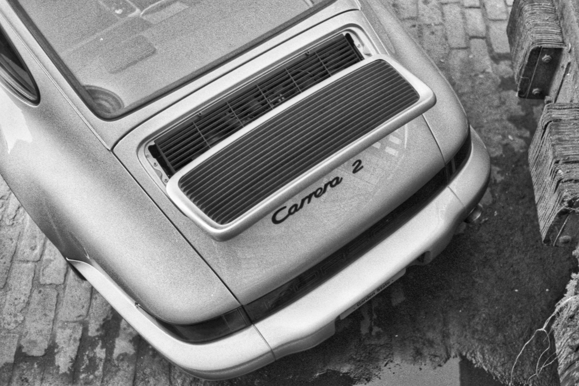 Black and white photo of the rear side of a vintage car, with a Toyota Carrera 2 badge, parked on a brick surface next to a wooden fence.