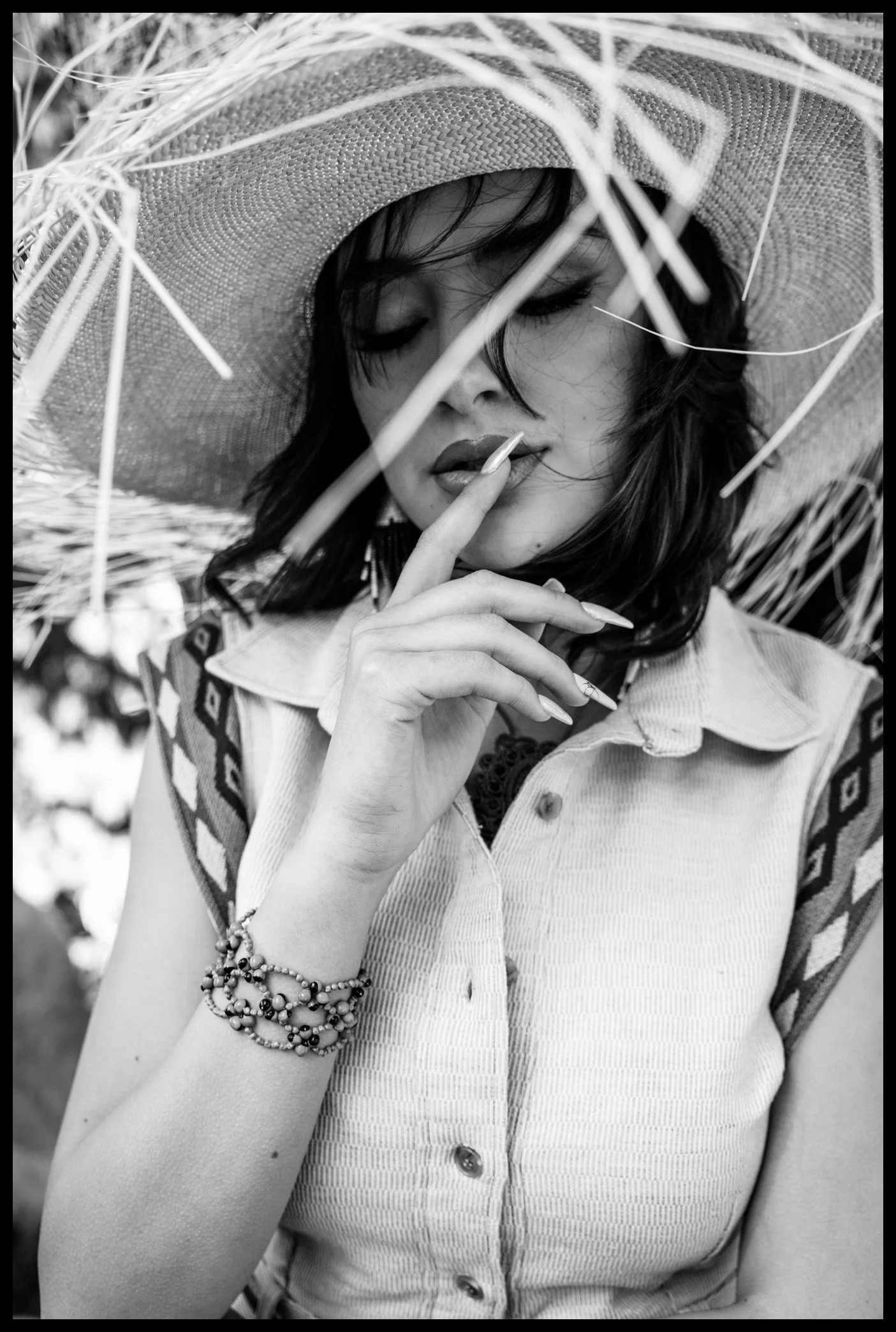 Black and white photo of a young woman wearing a large straw hat, with her eyes closed, touching her lips with her finger, wearing multiple beaded bracelets on her wrist, and dressed in a short-sleeved button-up shirt with a patterned detail on the s