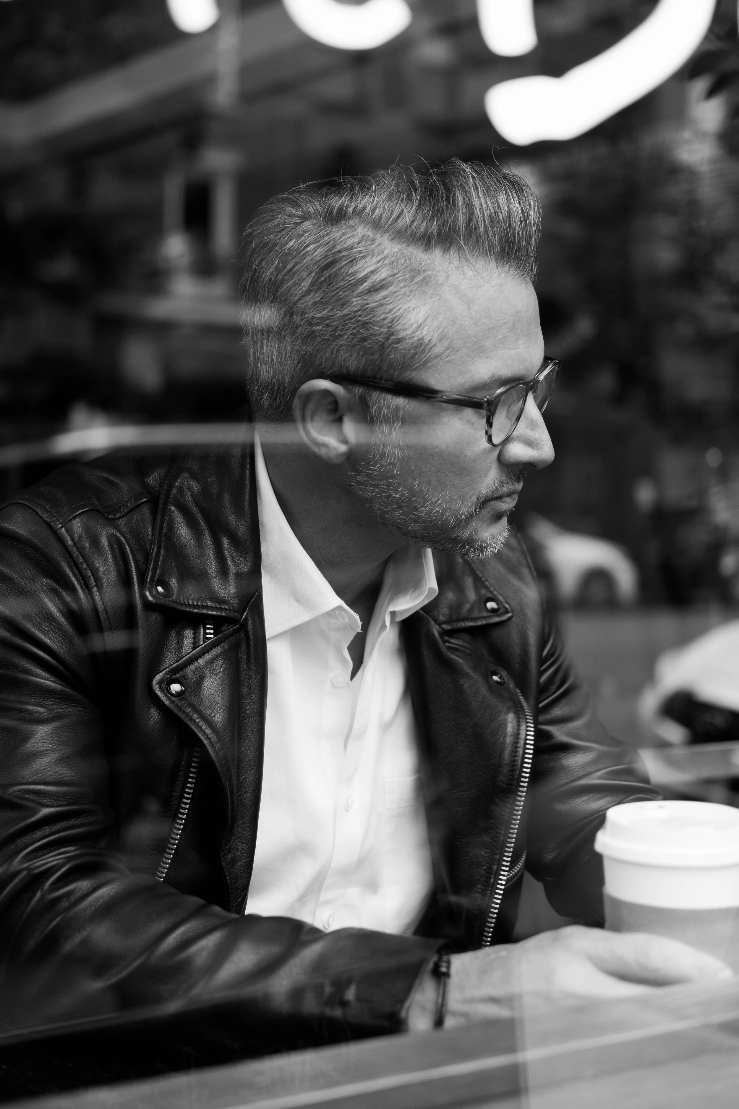 A man with glasses and gray hair sitting at a table, holding a coffee cup, wearing a white shirt and a leather jacket, inside a cafe or restaurant.