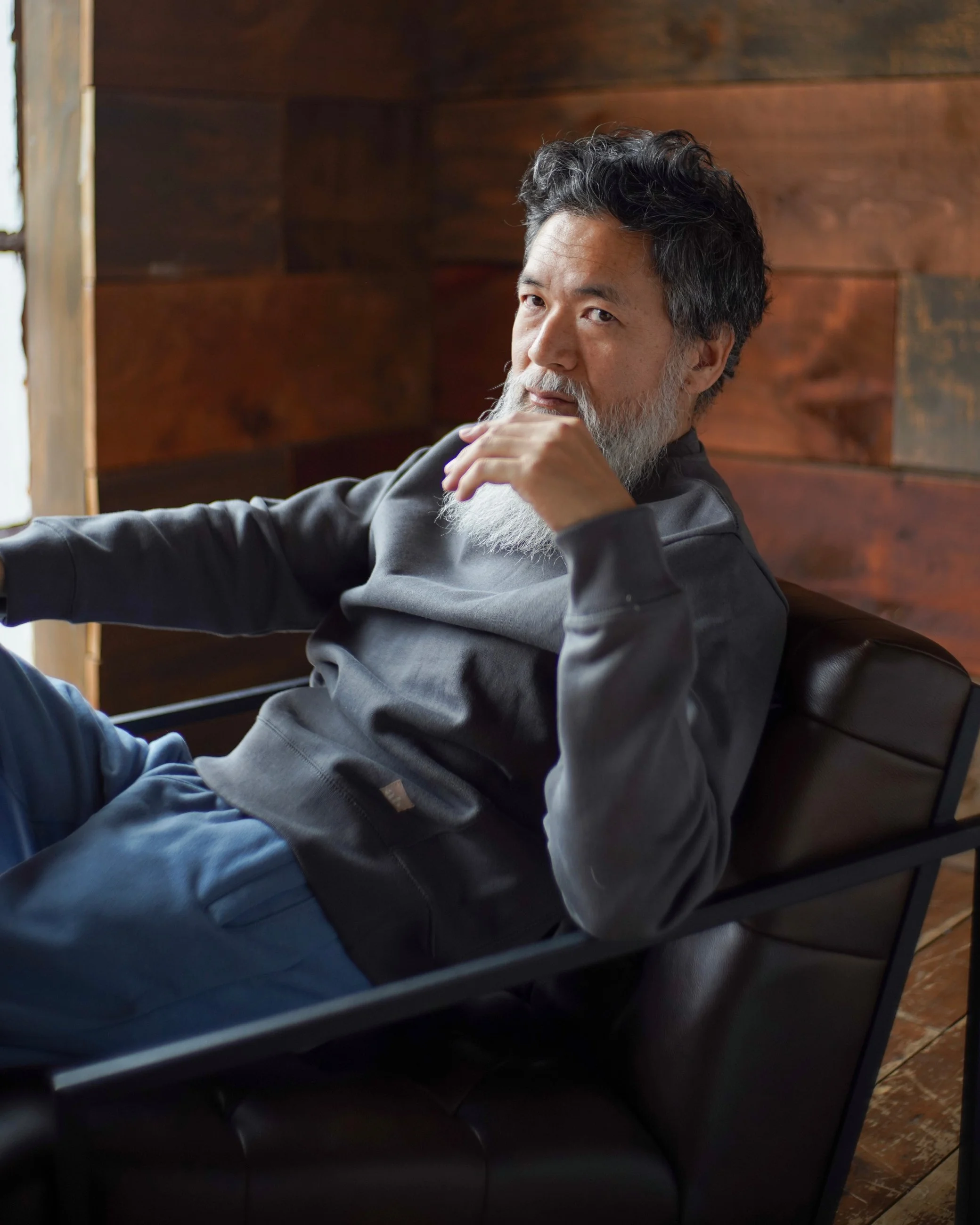 A middle-aged man with dark, curly hair and a gray beard, sitting on a black chair with a wooden wall in the background. He appears to be looking thoughtfully to the side, resting his chin on his hand.