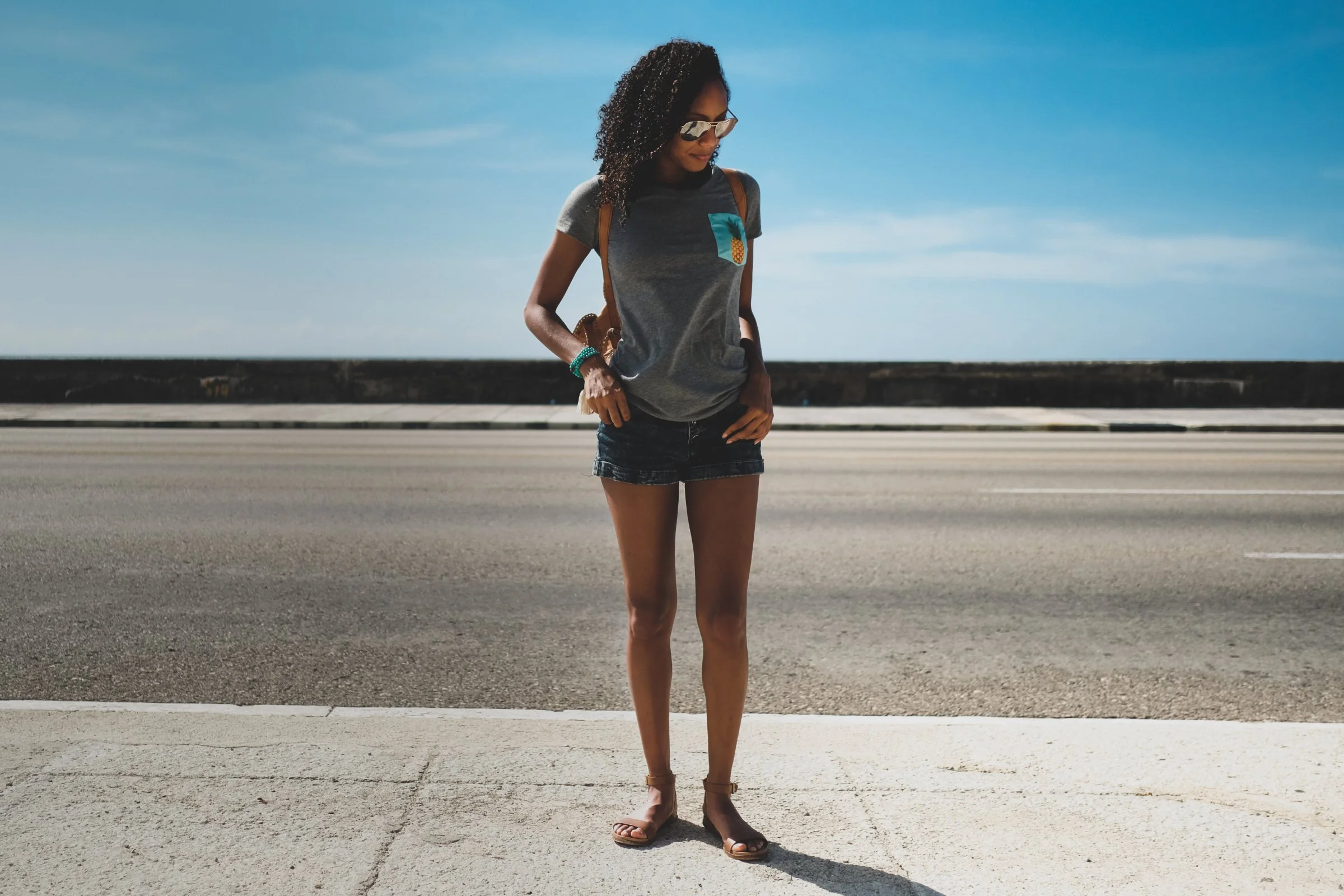 A woman in sunglasses, a gray t-shirt with a pineapple graphic pocket, denim shorts, and sandals walking by a beachside road under a blue sky.