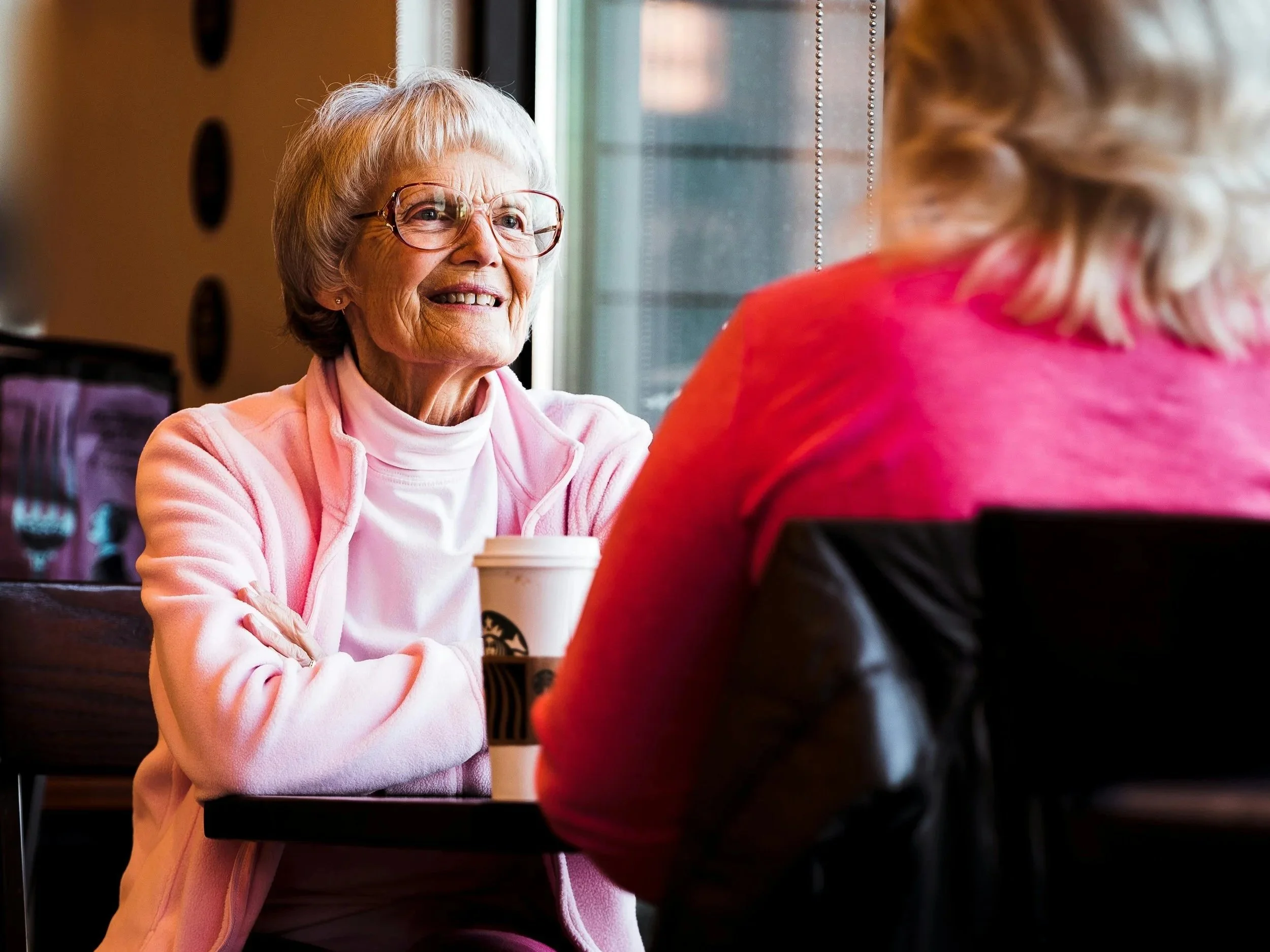 An elderly woman with glasses and a pink zip-up jacket smiling and talking to a younger woman with blonde hair in a pink shirt inside a coffee shop.