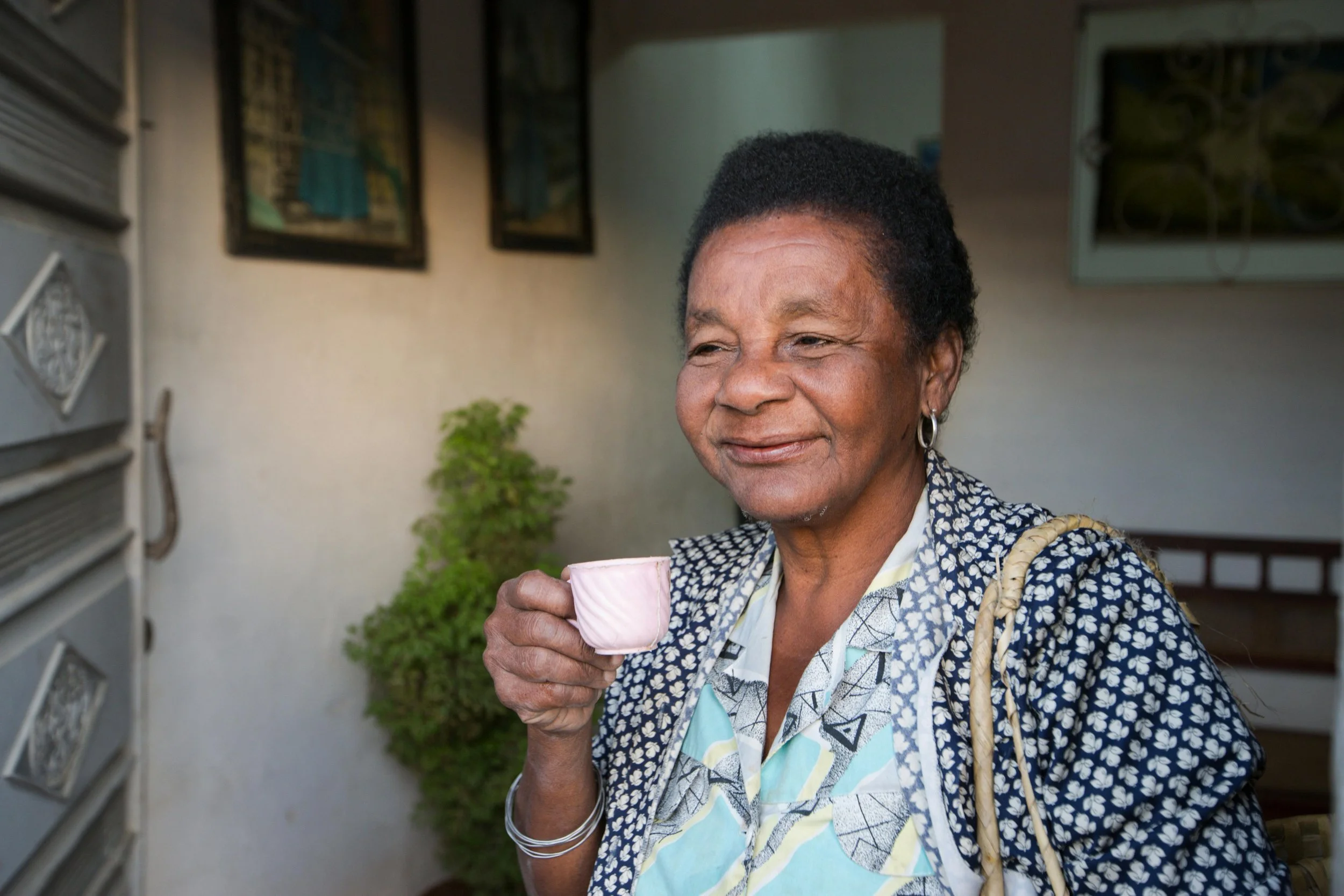 An elderly woman with short hair, wearing a patterned blouse and earrings, holding a pink cup and smiling peacefully outdoors.