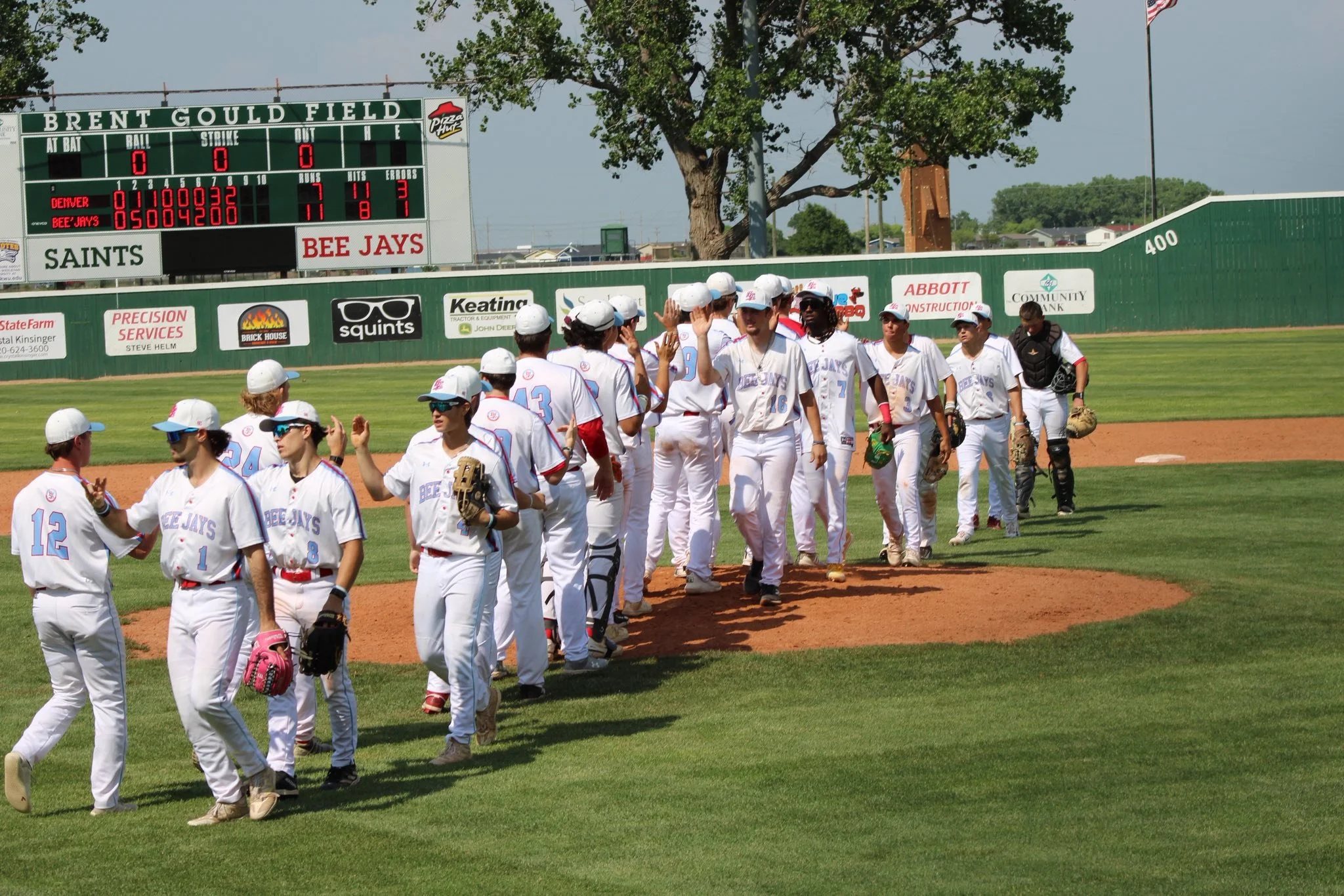 A group of softball players in white uniforms high-fiving each other on the field near a scoreboard at Brent Gould Field.