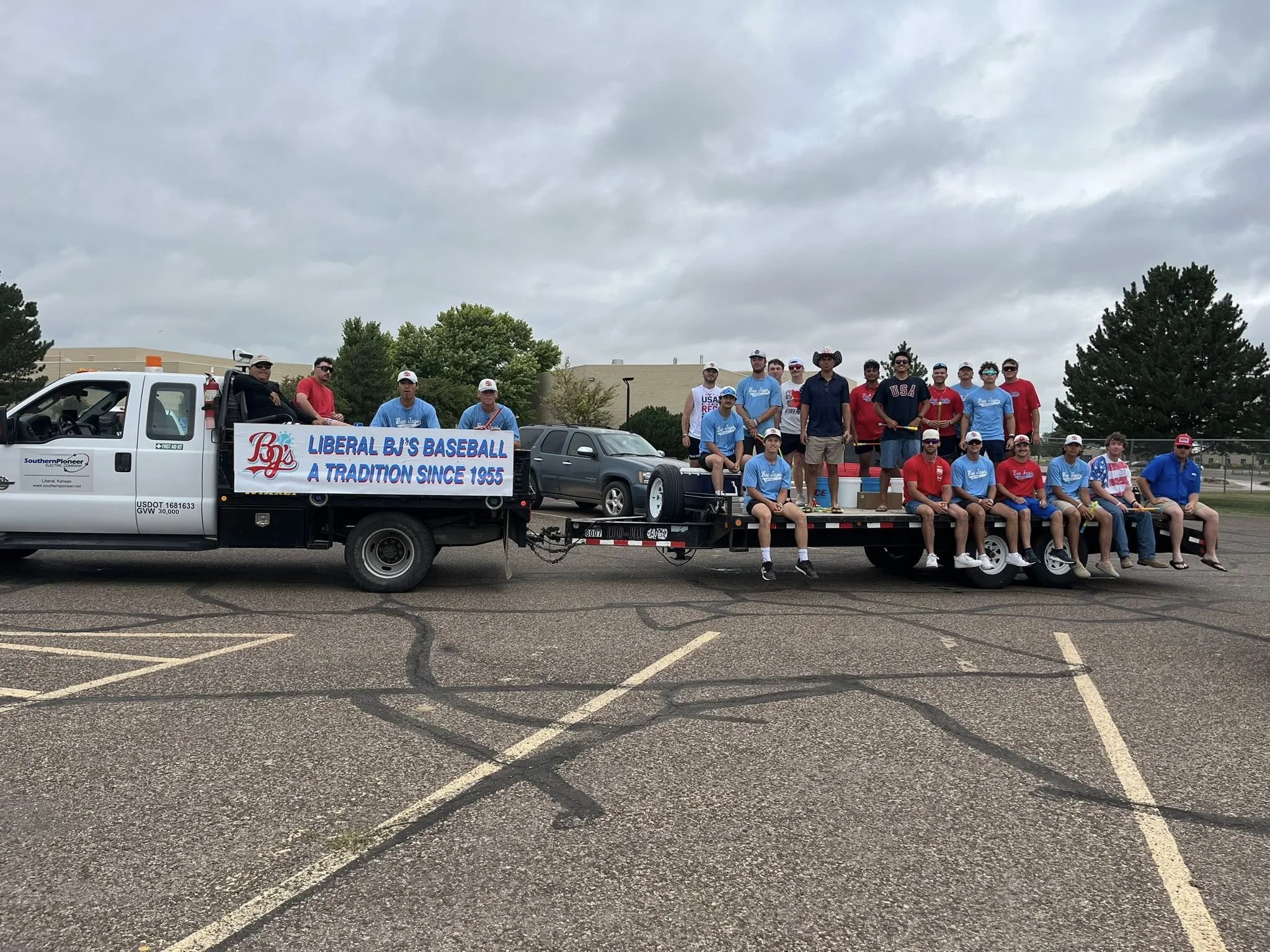 A group of young men and boys posing on a flatbed trailer with a sign that reads "Liberal BJ's Baseball - A Tradition Since 1955." They are outdoors on a cloudy day, with trees and a building in the background. Some are sitting, and others are standi
