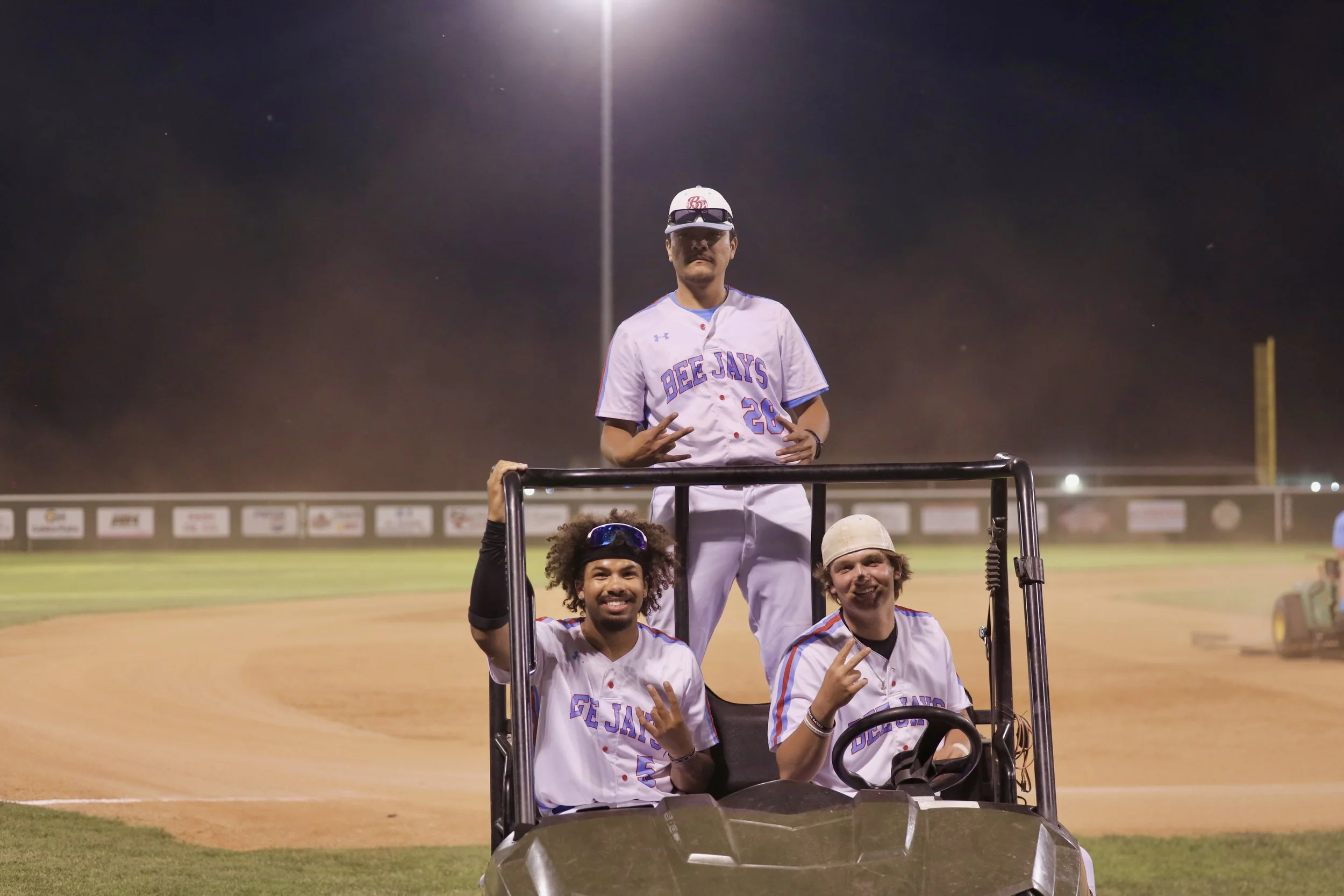 Three baseball players in uniform, two sitting in a golf cart and one standing behind it on a baseball field at night, with bright stadium lights above.