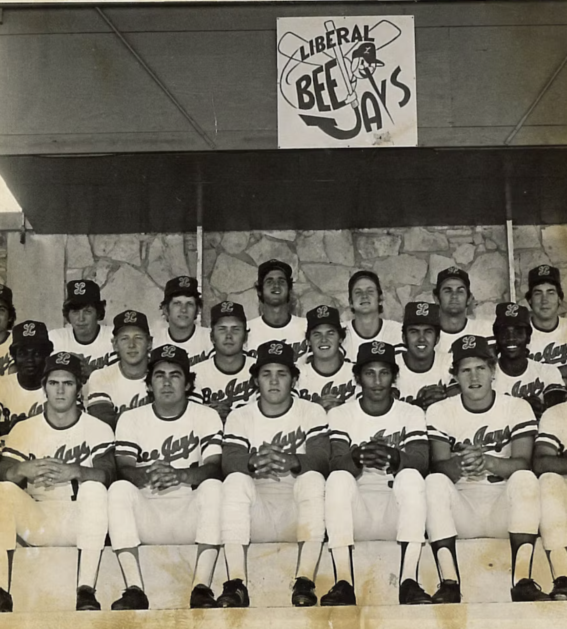 Black and white team photo of young baseball players wearing uniforms and caps, sitting and standing in front of a stone wall, with a sign above that reads 'Liberal Bee Jays'.