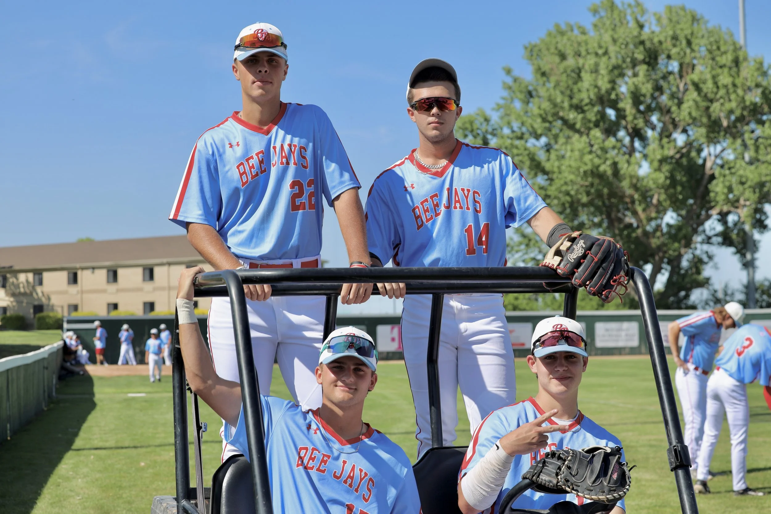 Two teenage boys wearing baseball uniforms and sunglasses sitting on a golf cart at a baseball field, with other players in the background.