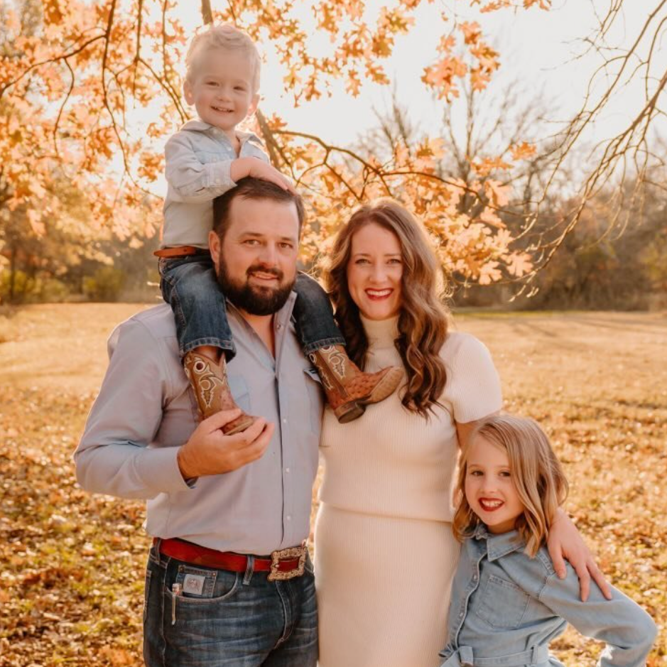 A family of four outdoors in a park during fall, with orange and yellow leaves on the trees. The father carries a young boy on his shoulders, and the mother stands beside him, smiling. A young girl stands in front, smiling and leaning against the mother.