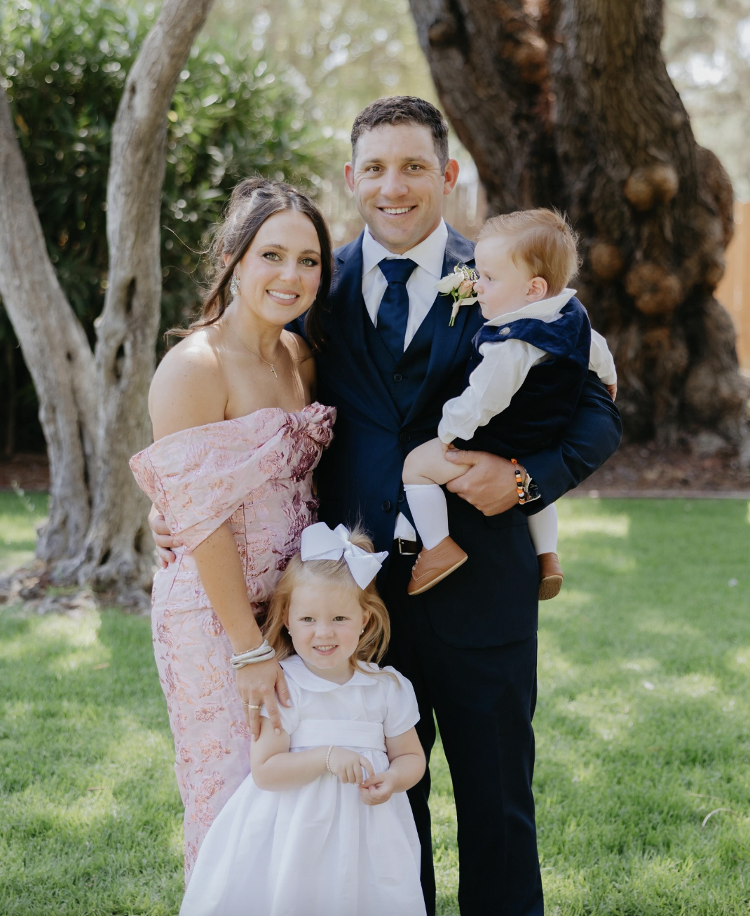 A family of four, including a man in a suit holding a young toddler boy, a woman in a pink dress, and a young girl in a white dress, smiling and standing outdoors in a park with trees.