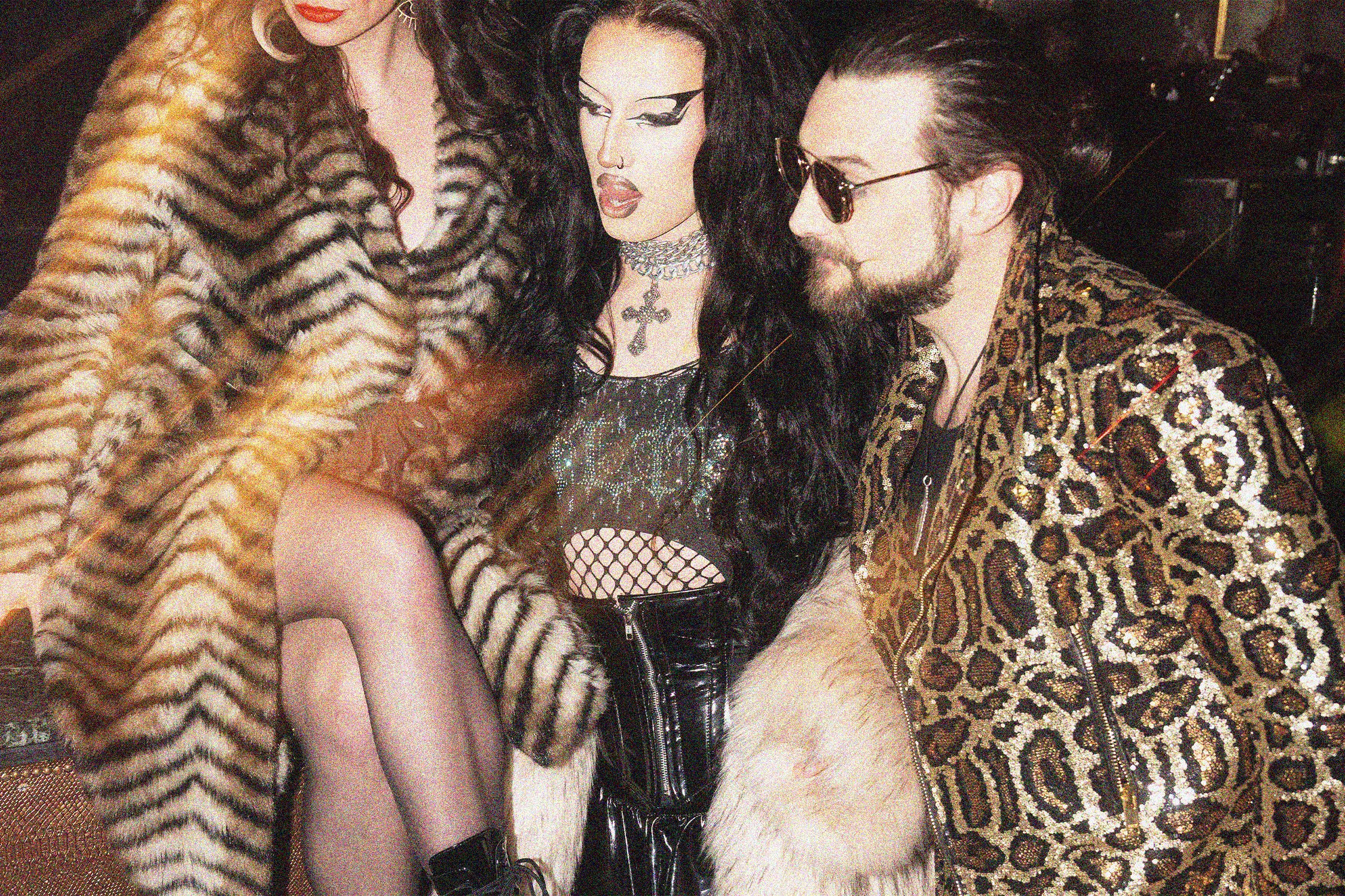 Three people dressed in glamorous clothing posing closely together, with leopard print coats and dark hair, in a social setting.