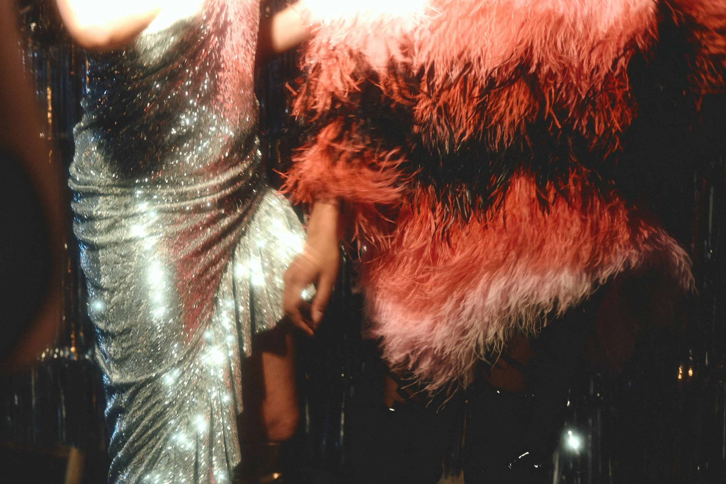 Close-up of two women holding hands, one in a shiny, silver, glittery dress, and the other in a vibrant, red, feathered outfit; night scene with small light reflections.