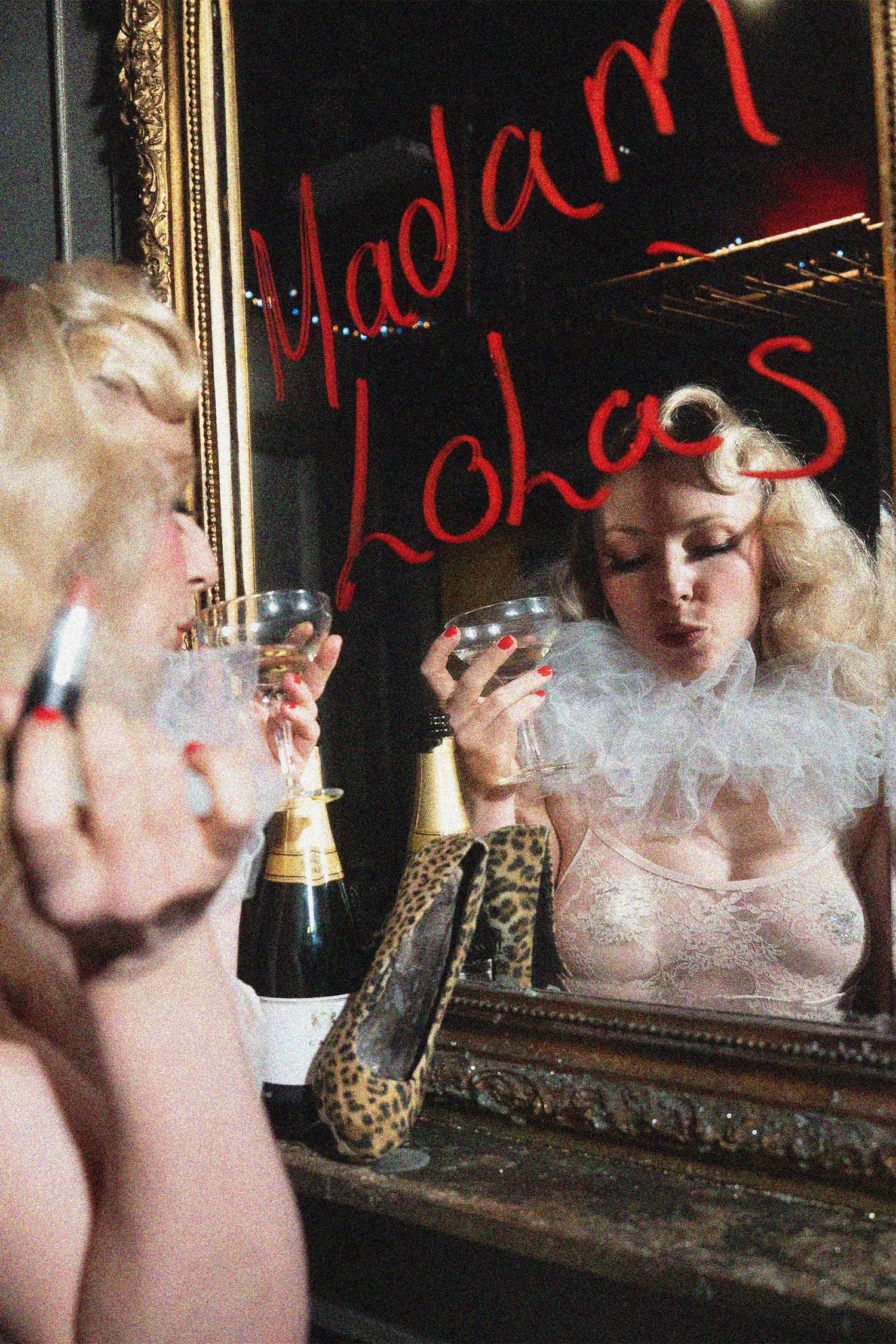A woman with blonde hair and red lipstick taking a photo with her phone in front of a mirror that has red cursive writing and a reflection of her face.
