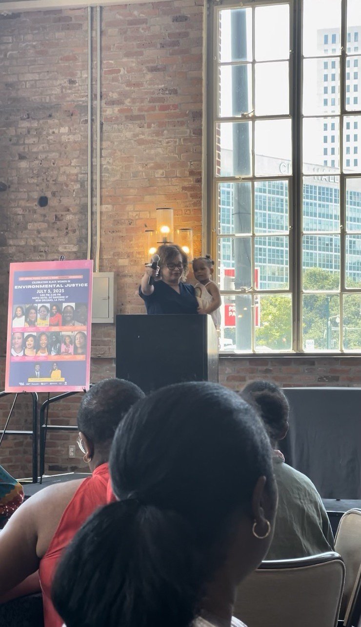 A woman holding a young girl standing at a podium during a public event in a room with large windows and exposed brick walls, with people seated in front of her.