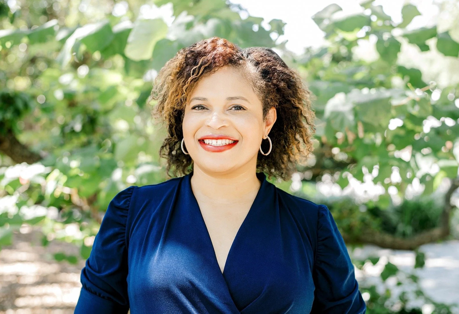 A woman with curly hair smiling outdoors, wearing a navy blue dress and hoop earrings, with green leafy background.
